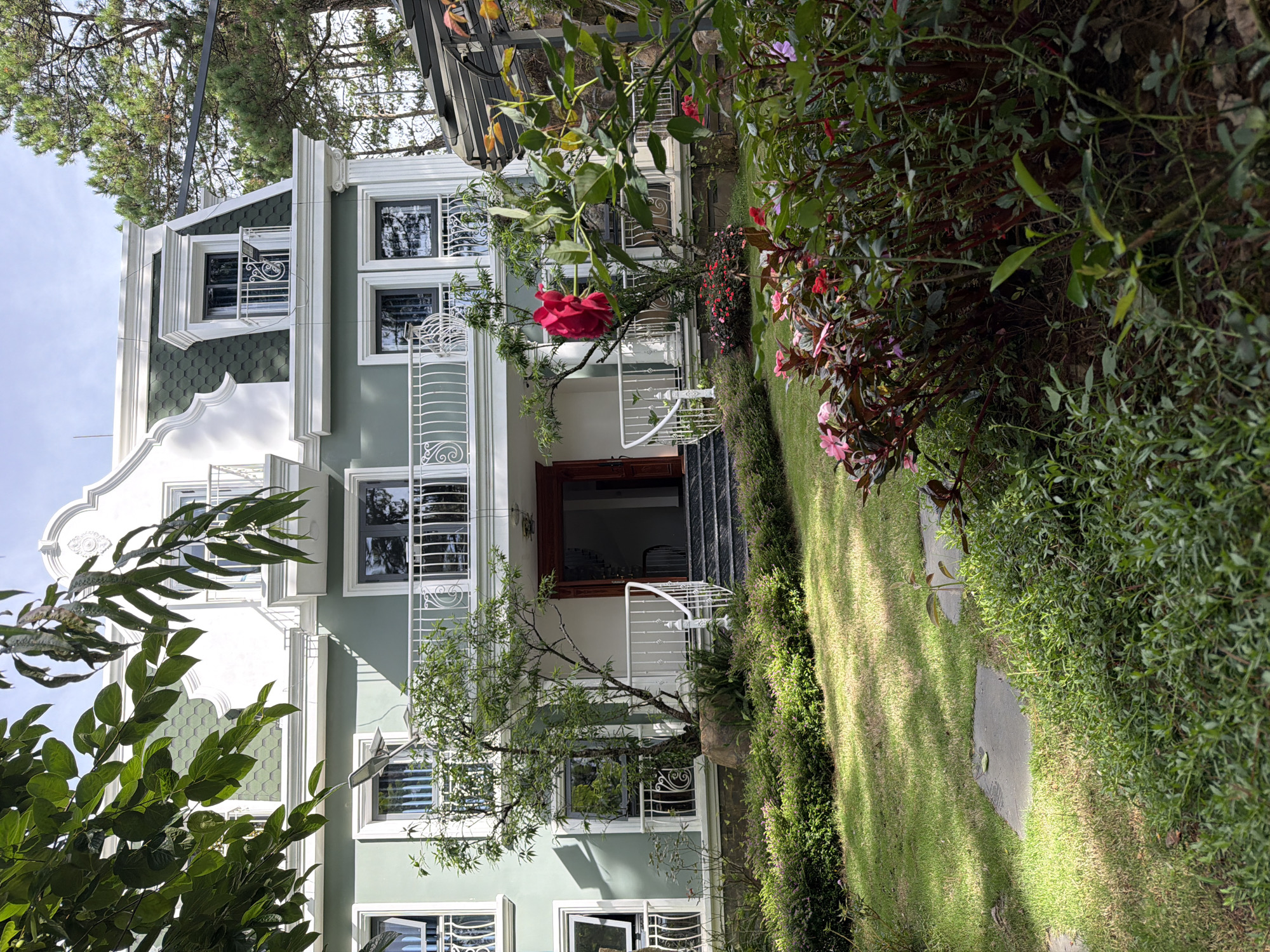 The image depicts the exterior of a multi-story house in Xuân Thành, Vietnam. The house has a light green facade with white trim and a dark green mansard roof. There are multiple windows and balconies with ornate white railings. A large tree with red flowers is prominently featured in front of the house, and lush green landscaping, including a manicured lawn and flowering bushes, surrounds the property. Pine trees are visible in the background. The scene is bathed in sunlight, casting shadows on the grass.