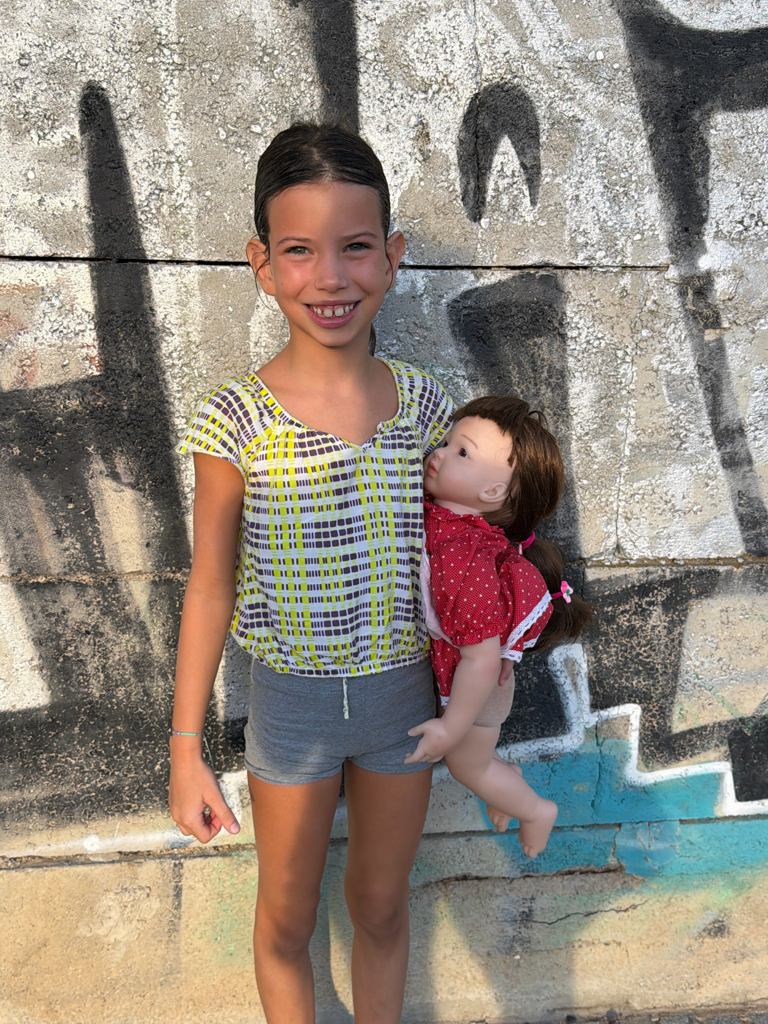 A young girl with dark hair, wearing a yellow and grey checkered shirt and grey shorts, stands in front of a graffiti-covered concrete wall. She is holding a doll with brown hair, wearing a red polka-dot dress. The girl is smiling and looking at the camera. The concrete wall appears weathered and has sections of dark and light gray paint, with prominent black graffiti and some blue paint. The ground in front of the wall is also concrete, with shadows cast by the girl and the wall's graffiti. The image seems to be taken outdoors, in a public urban space. The location is identified as Salou, Spain.