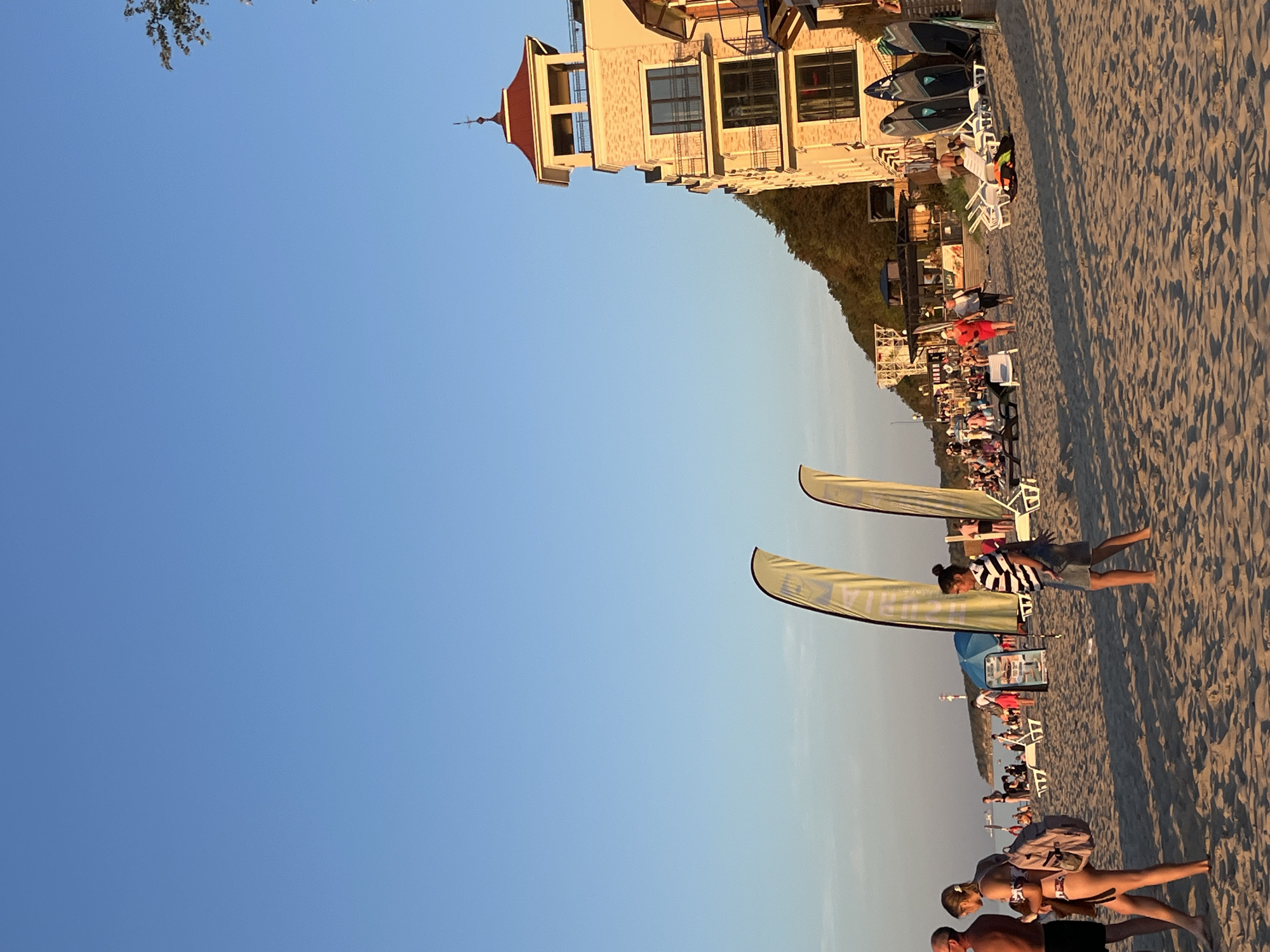 This image captures a lively scene on a sandy beach in Pionersky, Russia, during what appears to be late afternoon or early evening, indicated by the warm, golden light. The sky is a clear, vibrant blue.
In the foreground, several people are walking on the sand. On the left, a man with a backpack is seen from the side. To his right, a woman in a bikini and a backpack is walking away from the viewer, also seen from the side. Further to the right, a person wearing a striped t-shirt and denim shorts is walking towards the right of the frame.
Scattered across the beach are numerous beach chairs, some occupied, suggesting a relaxed atmosphere. In the middle ground, two large, yellow, feather-shaped flags stand prominently. Beyond them, a crowd of people are visible, some sitting and some standing, enjoying the beach. A small, blue umbrella is also present in this area.
To the right side of the image, a multi-story building with a distinctive red-tiled roof and balconies dominates the scene. The building's facade is light-colored, with a pattern of darker bricks. In front of the building, several surfboards or paddleboards are propped up vertically. Further back, more buildings and structures can be seen along the coastline, nestled against a densely forested hillside. A lighthouse or similar structure is visible in the distance on the left. The overall impression is of a popular tourist destination with various activities and amenities.