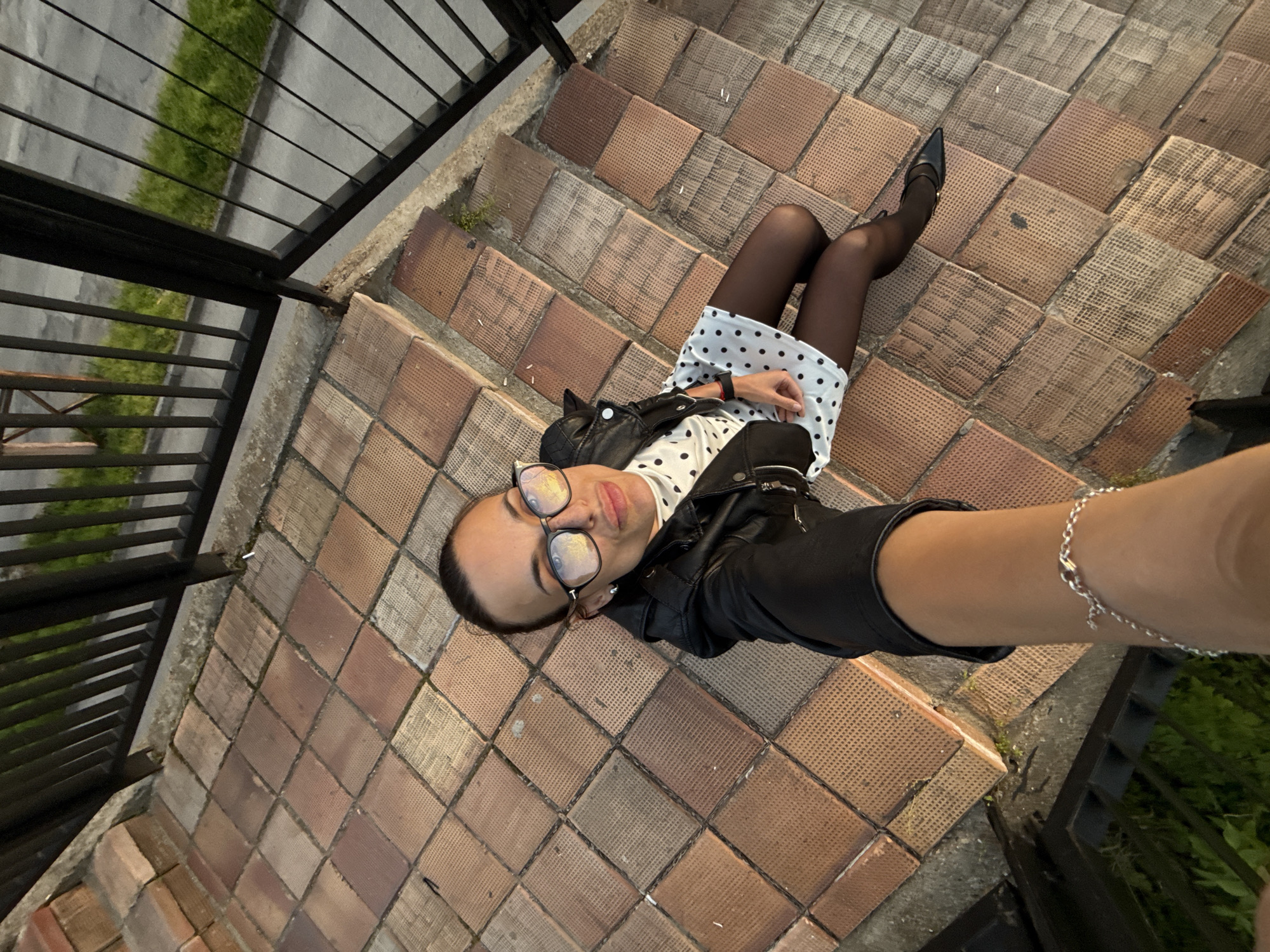 A woman is depicted lying on her back on a set of outdoor stairs in Pleven, Bulgaria, looking directly up at the camera for an overhead selfie. She has dark hair pulled back and is wearing clear-lensed glasses, a black leather jacket, a white dress with black polka dots, black sheer tights, and black pointed-toe shoes. A silver chain bracelet adorns her right wrist.
The stairs are composed of square, textured tiles in varying shades of reddish-brown and gray. Black metal railings with horizontal bars border both sides of the stairway. Beyond the railings, patches of green grass and a gray concrete or paved surface are visible. The lighting suggests it is either late afternoon or early evening. No text is visible in the image.