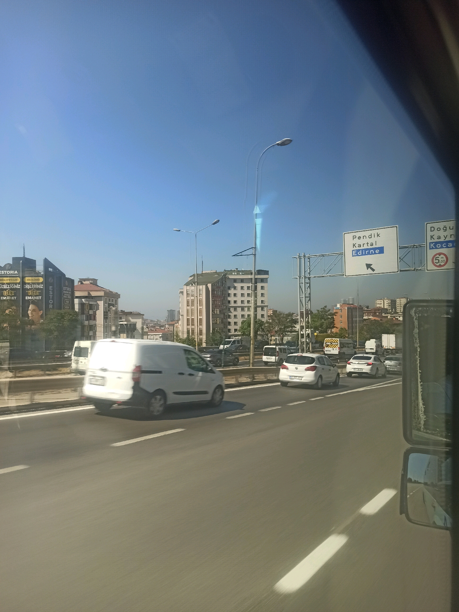 The image captures a bright, sunny day in Pendik, Turkey, with a view from a vehicle's perspective. The road ahead shows several cars in motion, including a white van in the foreground and two white sedans further down the highway. Overhead, a large road sign indicates directions to "Pendik," "Kartal," and "Edirne." Another sign displays "Doğu," "Kayn," and "Koca" with a speed limit of 50 km/h. In the background, numerous buildings of varying heights form a cityscape, with a few trees interspersed. The sky is a clear, vibrant blue. The presence of the road signs and the urban architecture suggests a highway in a developed area of Pendik.