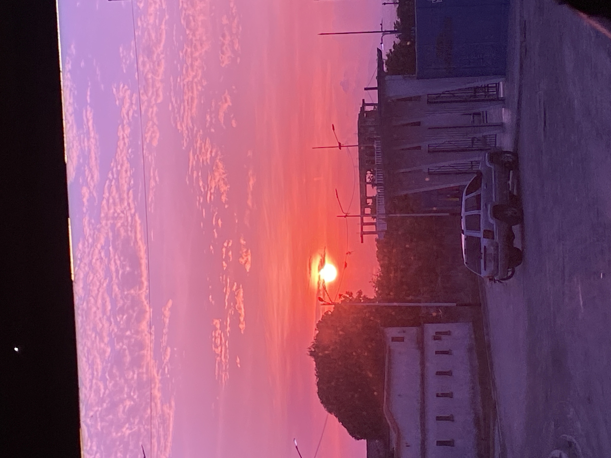 The image depicts a sunset in Higuerote, Venezuela. The sky is a gradient of pink and purple hues with scattered white clouds. In the foreground, there is a street scene with buildings, trees, streetlights, and a white van parked on the right side. The sun is setting behind the trees, creating a bright spot in the center of the image.