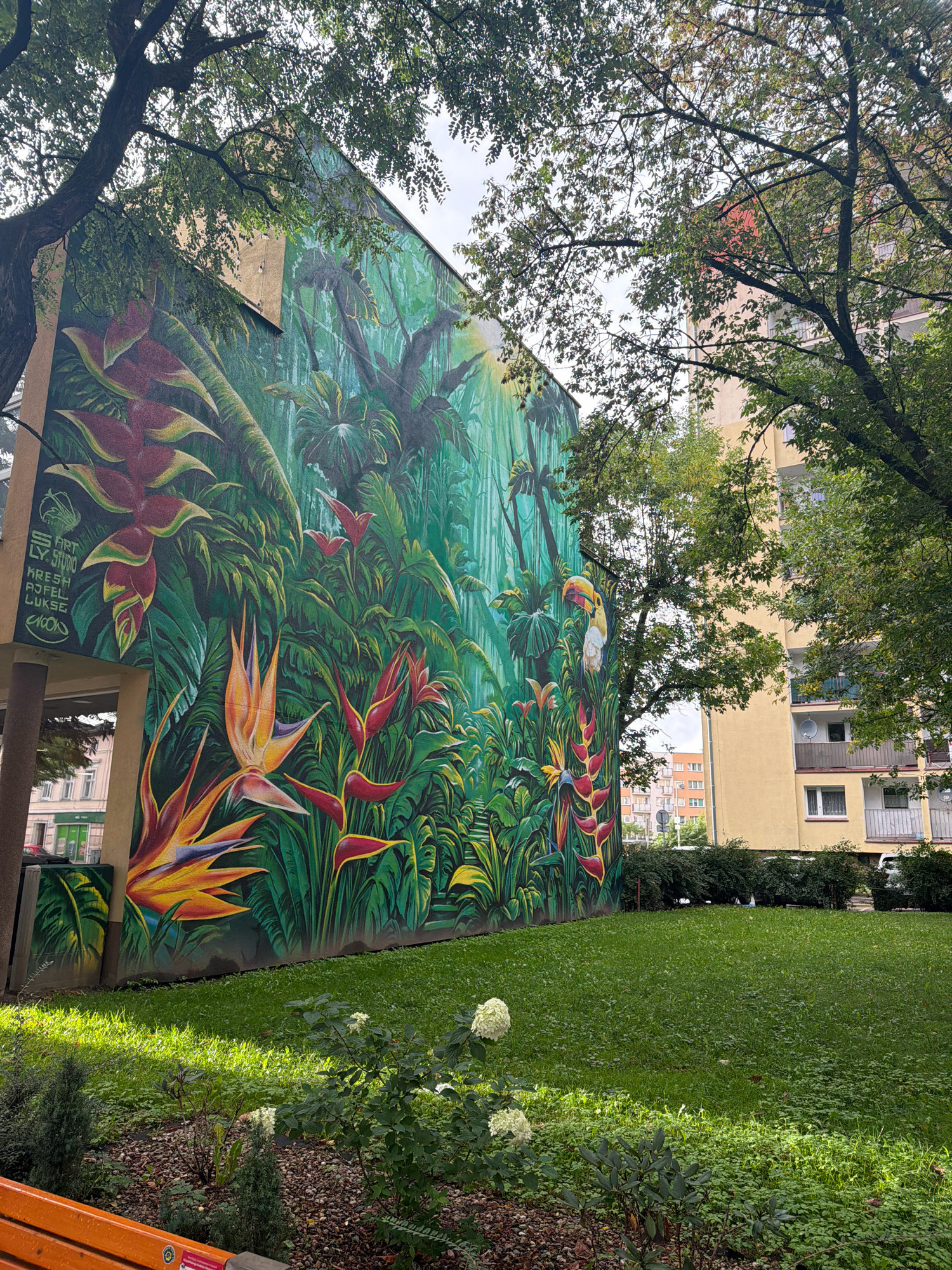 The image depicts a vibrant mural of a tropical rainforest scene painted on the side of a building in Koszalin, Poland. The mural features lush green foliage, large red and yellow flowers resembling Heliconia and Bird of Paradise plants, and a toucan perched amidst the leaves. The building itself appears to be an apartment complex, with a lighter colored facade and balconies visible in the background. In the foreground, a well-maintained lawn is present, with some small bushes and a portion of an orange bench visible at the bottom left. The mural is an artistic rendition and does not depict any actual animals or people in the scene. The overall impression is one of urban art adding a splash of nature to the cityscape.