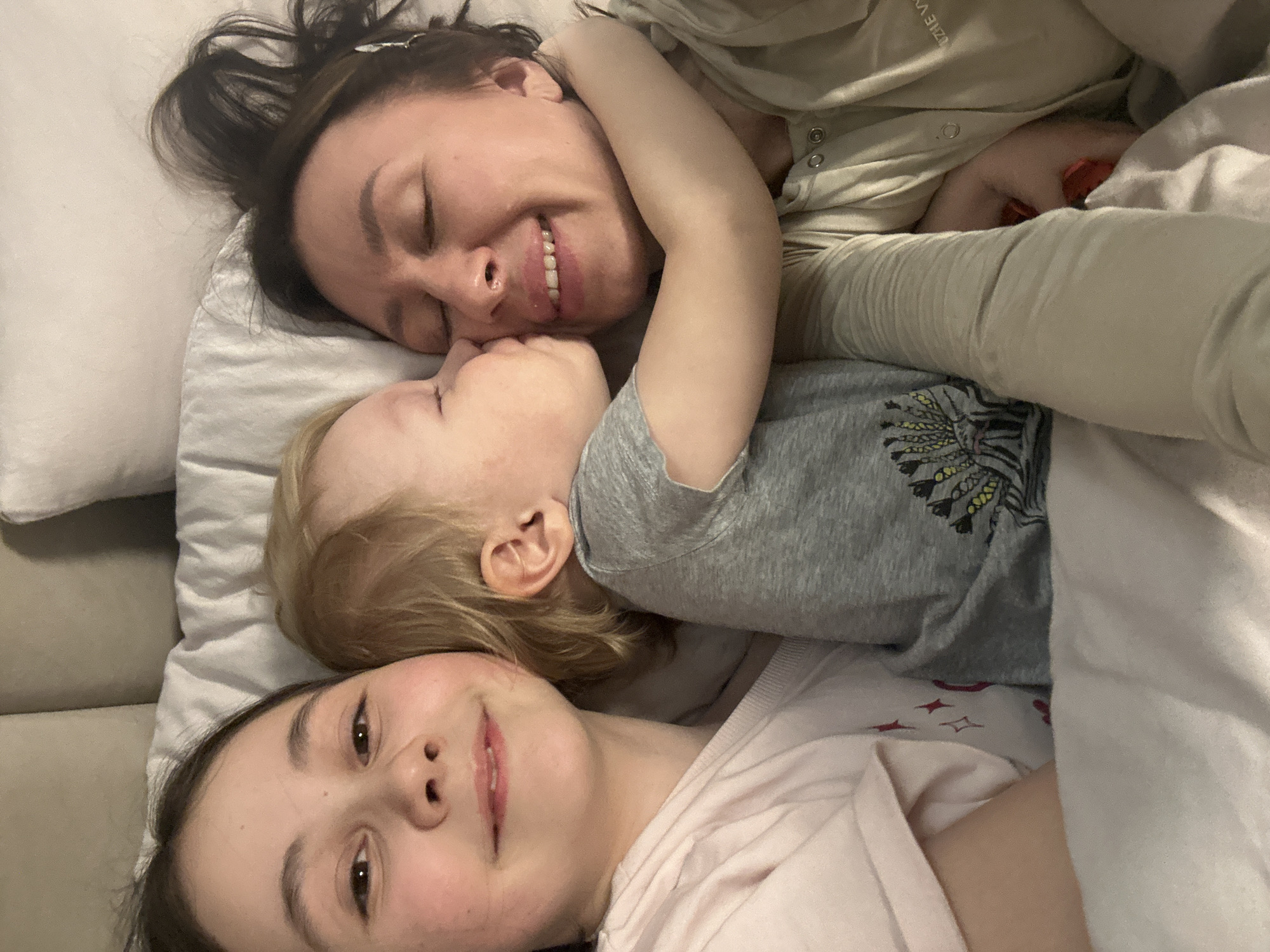 Three individuals, an adult woman and two children, are captured in a close-up, top-down shot, lying together in an intimate indoor setting. The adult, positioned at the top of the frame, has dark hair, closed eyes, and a joyful smile, wearing a light-colored long-sleeved top with faint, unidentifiable text. Nestled against her face is a young child with blonde hair and closed eyes, wearing a grey t-shirt adorned with a stylized animal or feathered graphic. At the bottom of the frame, an older child with dark hair looks directly at the camera with a gentle smile and open eyes, dressed in a light pink top.
They appear to be relaxing or cuddling on a bed or sofa, surrounded by soft pillows and blankets, with a light-colored sofa visible on the left. The scene evokes feelings of warmth, love, and contentment, suggesting a peaceful moment shared between a family, possibly a mother and her children. The lighting is soft, indicative of morning or evening. This tender family moment takes place in Votkinsk, Russia.