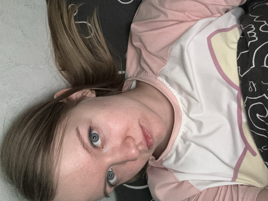 The image presents a close-up portrait of a single female individual in an indoor environment within Nizhnekamsk, Russia. The subject has fair skin with a slight reddish tone, light blue eyes, and light brown hair. She looks directly into the camera with a neutral to slightly somber expression. Her hair is partially visible, extending towards the right side of the frame, and a small gold-colored hoop earring is present in her right earlobe. She is wearing a long-sleeved top with a white torso and light pink raglan sleeves; the white section displays a light yellow and pink graphic design near the lower edge. The individual is positioned in a reclining manner. A dark grey fabric with white abstract patterns is visible around her, functioning as both a bedding item and a background element. Behind her head, a textured, light grey wall is visible. No other individuals, significant objects, or specific actions beyond posing for the photograph are discernible.