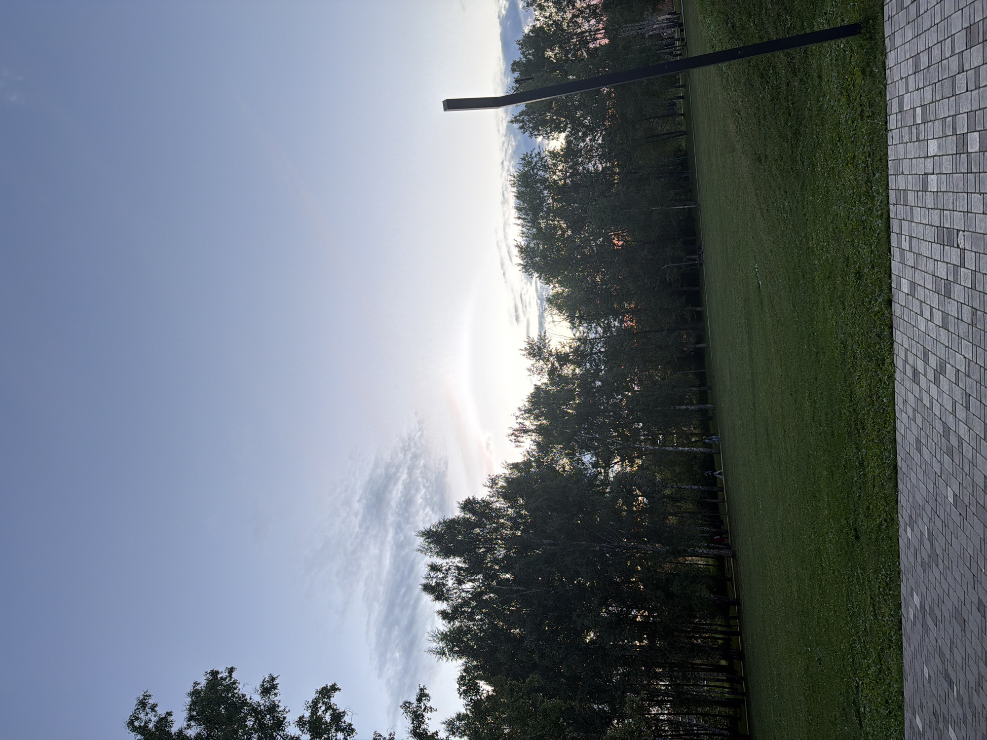 The image depicts an outdoor scene, likely in Nizhnekamsk, Russia, during what appears to be late afternoon or early evening given the soft, diffused light. The sky dominates the left side of the frame, a clear, pale blue transitioning to a brighter, almost hazy horizon where the sun is likely setting or has recently set. Wispy clouds are scattered across this brighter area, catching the light and adding texture.
A line of dark green trees runs horizontally across the middle of the image, their dense foliage silhouetted against the sky. Beyond the trees, a glimpse of what might be buildings is visible, suggesting an urban or park-like setting within the city. A prominent, dark, angular object, possibly a sculpture or architectural element, juts out from the top of the frame and extends downwards, partially obscured by the trees.
To the right of the trees, a neatly mowed expanse of green grass stretches out, suggesting a park or a well-maintained lawn. The right edge of the image is bordered by a textured surface composed of small, grey rectangular tiles, arranged in a grid pattern, possibly a wall or pathway.
There are no visible people or text in the image. The overall mood is serene and tranquil, with the natural elements of the sky and trees contrasting with the man-made tiled surface. The lighting and the long shadows cast by the trees would indicate it's either sunrise or sunset.