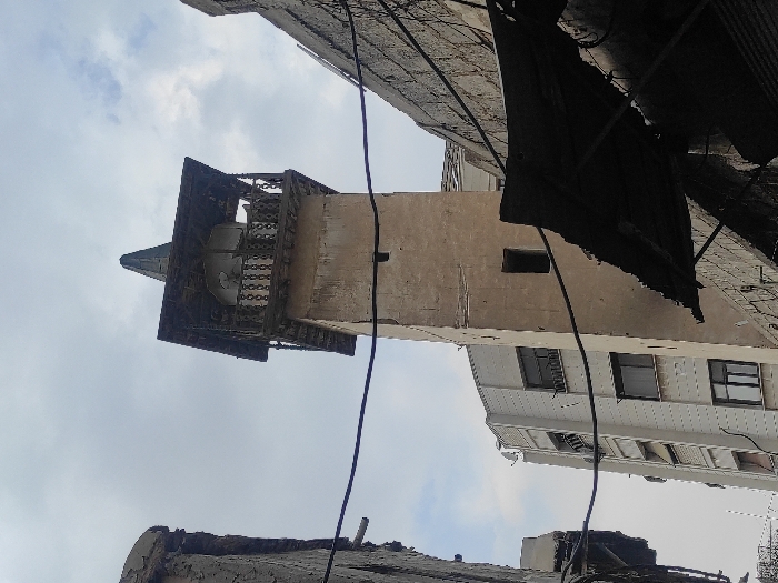 The image displays an urban scene within Al-Qanawat Municipality, Syria. A weathered, light-brown minaret with a square shaft and a single rectangular opening dominates the central view. The minaret's summit features a wooden balcony with a balustrade, topped by a dark, pointed, pyramidal roof. Two loudspeakers are discernible within the balcony structure. To the left and rear of the minaret, a taller, modern, light-colored multi-story building with multiple dark-framed windows and balconies is visible. On the right side, dark, tattered fabric shades are suspended above a structure, partially obscuring a rough-textured wall. A damaged wall segment is present on the far left. Multiple black electrical or communication wires crisscross the frame against an overcast sky with visible clouds. No individuals or specific actions are depicted.