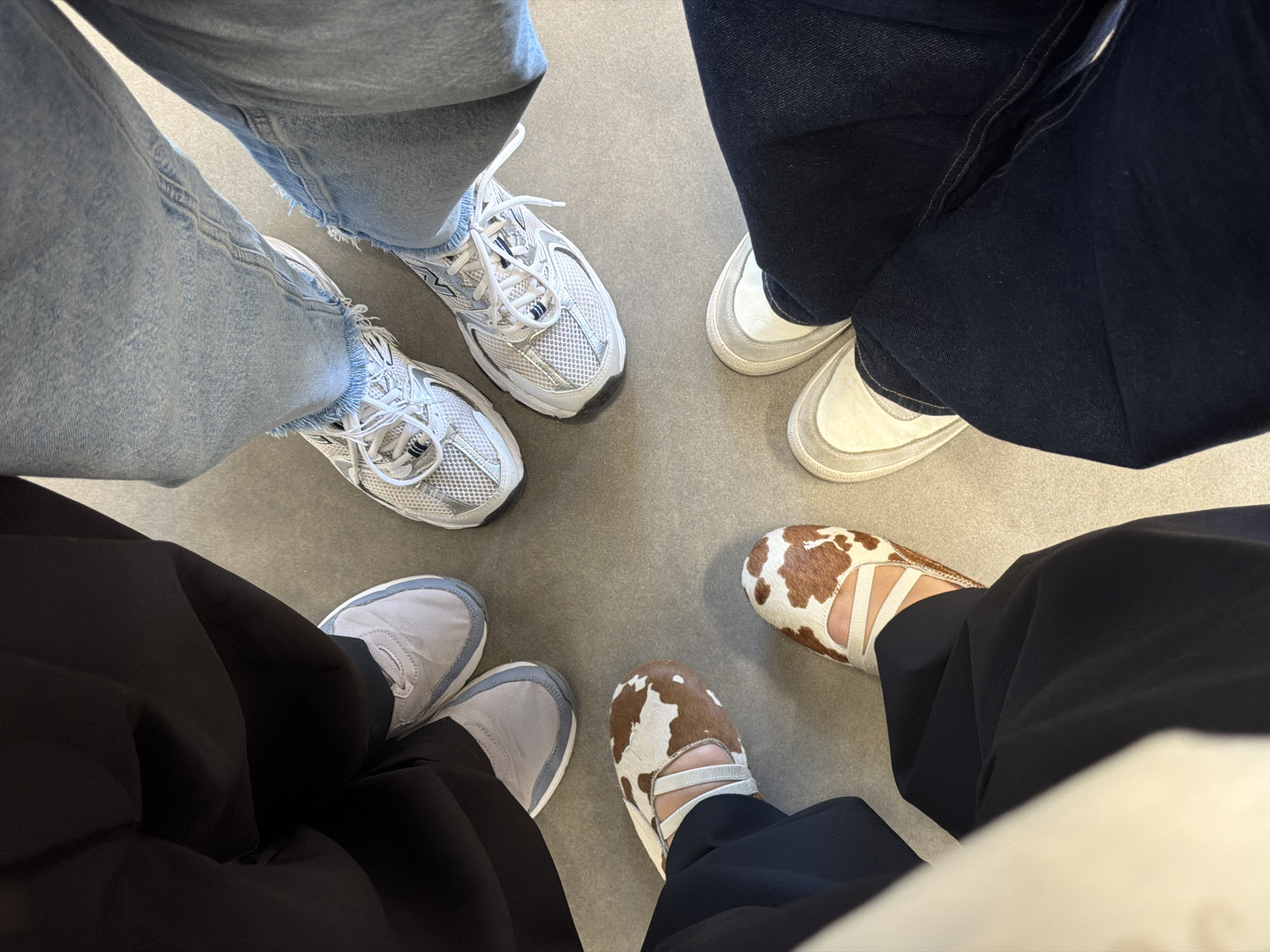 The image depicts a group of five people standing in a circle, showing only their legs and shoes. The background is a plain grey floor. The individuals are wearing a variety of footwear: two wear white athletic shoes with light blue jeans, one wears dark denim jeans with white sneakers, one wears black pants with gray sneakers, and another wears black pants with cow-print Mary Jane shoes. The image does not provide enough information to determine the location as Yerevan, Armenia.
