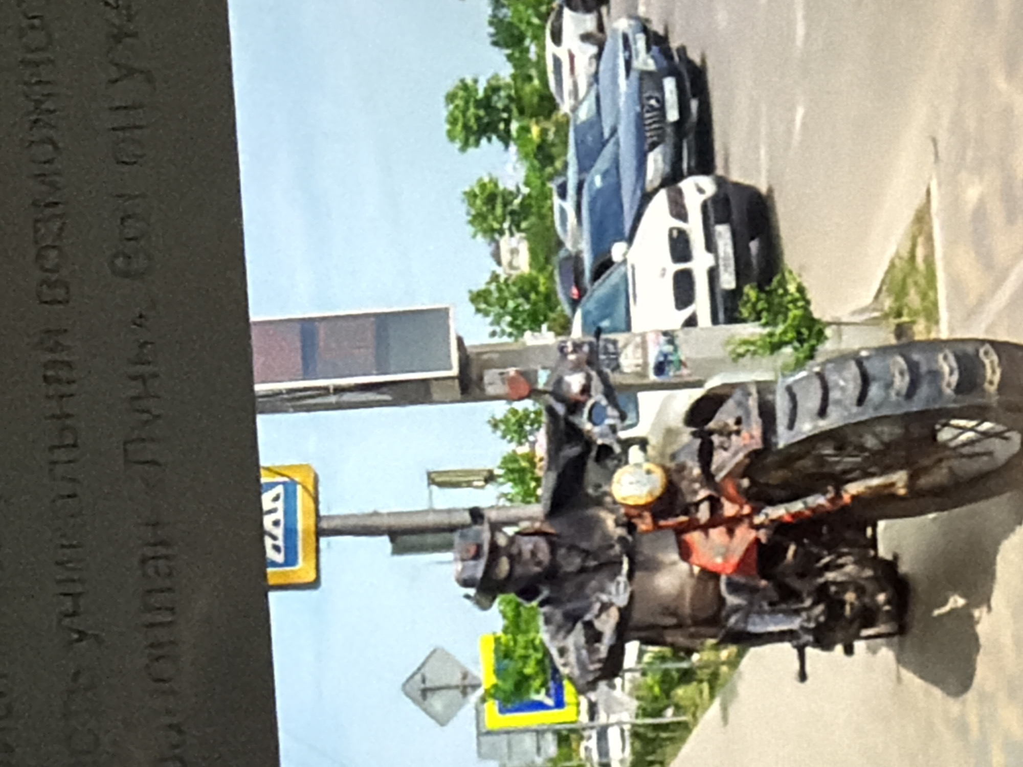 A weathered statue of a person in a hat and goggles rides a motorcycle on a city street. The statue is dark and appears to be made of metal. Behind the statue, several cars are parked along the road, and a pedestrian crossing sign is visible. The sky is blue, and trees line the background. The statue's attire suggests a vintage or steampunk aesthetic.