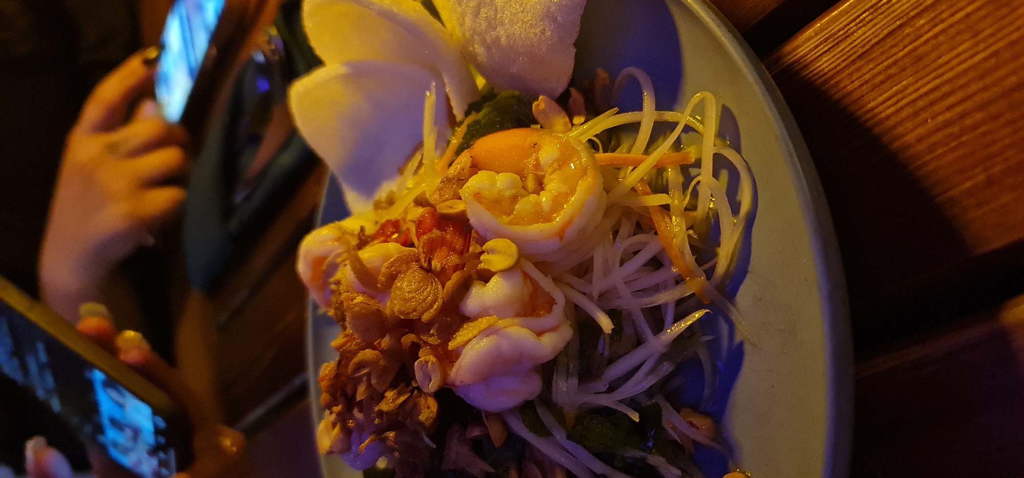 A dimly lit, close-up shot of a plate of Vietnamese food, likely Mi Quang, sits on a wooden table in a restaurant. The plate is blue and holds a vibrant dish consisting of prawns, rice noodles, shredded carrots, herbs, fried garlic, peanuts, and rice crackers. The prawns are cooked and have a distinct orange color. A person is partially visible in the background, holding a smartphone and focusing on the food, possibly taking a picture. The scene suggests a casual dining setting. It is not possible to confirm the location is Đà Nẵng, Vietnam with the current information.
