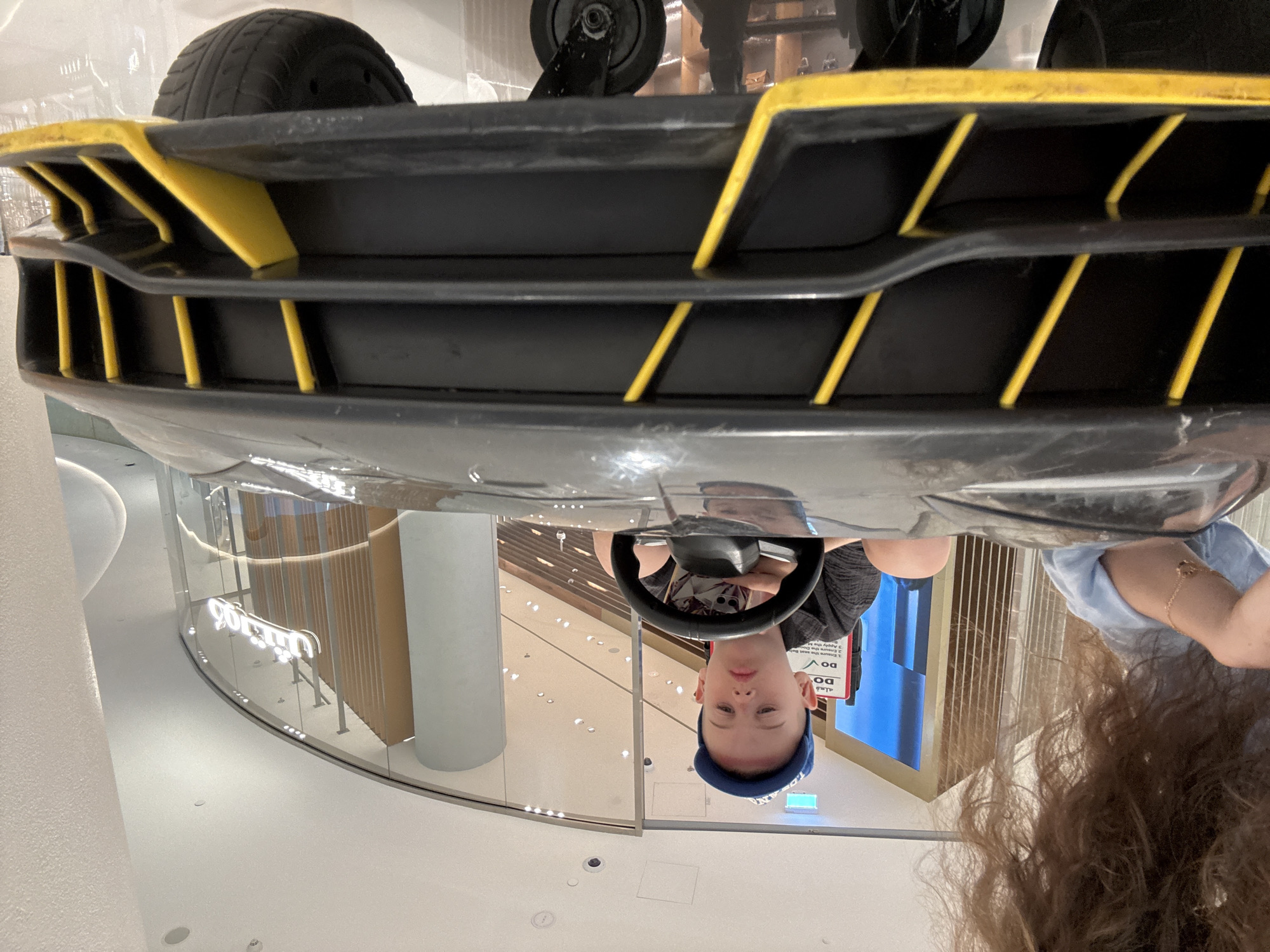 A young Caucasian boy wearing a blue baseball cap and a dark grey t-shirt sits in the driver's seat of a black toy car with yellow accents. He is holding the steering wheel and looking forward with a neutral expression. The car appears to be in a shopping mall, as indicated by the polished floor, modern architecture, and signage in the background, including Arabic script on a glass storefront. A woman's shoulder and long brown curly hair are visible on the left side of the frame, suggesting she is with the boy. The car has a low profile with a wide front bumper featuring yellow vertical slats. The environment is brightly lit, with a high ceiling and circular light fixtures.
