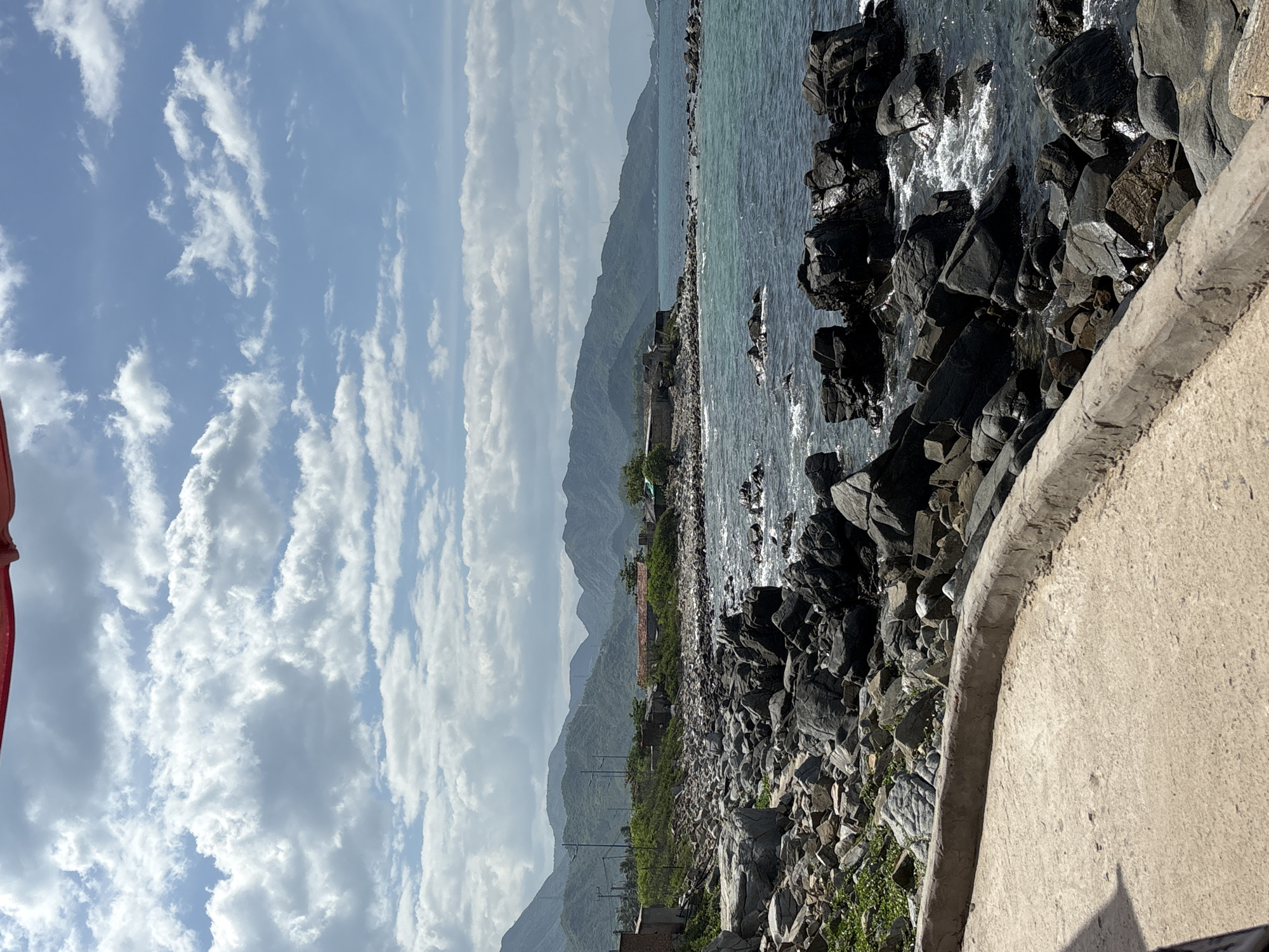 The image depicts a rocky coastline in Vĩnh Lương, Vietnam. A concrete path curves into the foreground, bordered by a low concrete wall. Beyond the path, a dense collection of dark, weathered rocks forms the immediate shoreline. The sea is visible to the right, with waves gently crashing against the rocks, creating white foam. In the mid-ground, more rocks lead to the water's edge, with some sparse green vegetation growing among them. In the distance, the coastline is lined with low buildings, some with terracotta roofs, and lush green hills rise behind them. The sky is a bright blue, filled with scattered white clouds. The overall scene is one of a natural coastal landscape with some signs of human habitation.