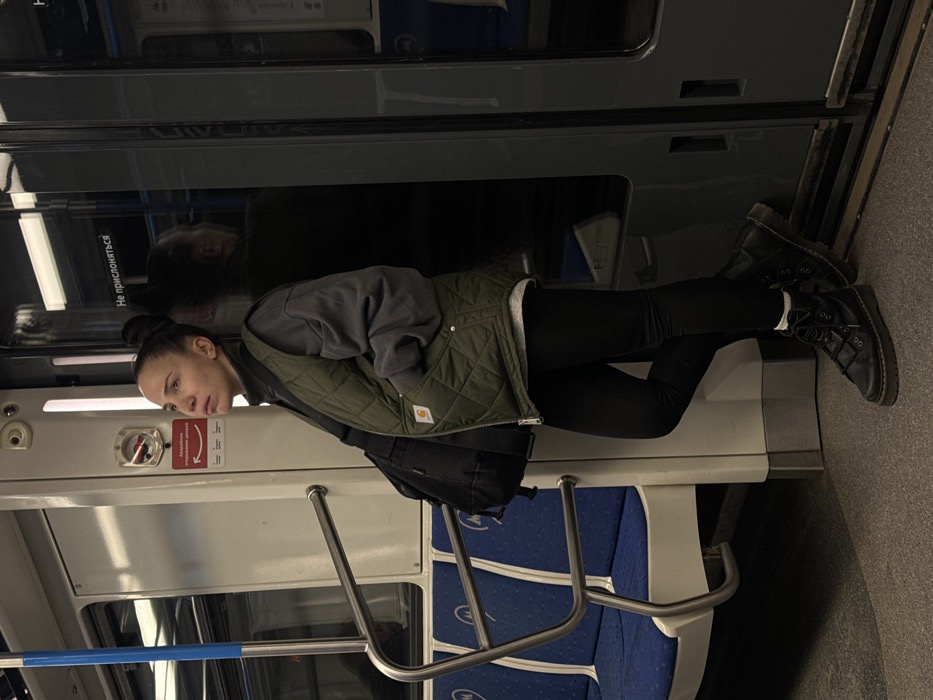 A single female subject stands inside a Moscow Metro subway car. She is positioned towards the right side of the frame, leaning against a dark gray doorframe with her right leg bent and her left foot flat on the gray floor. Her hands are placed into the pockets of her olive green quilted Carhartt vest, worn over a gray long-sleeved sweatshirt and a black turtleneck. She wears black leggings and black combat boots with visible white socks. A black backpack is slung over her left shoulder. Her dark hair is pulled back into a bun, and she looks upward and slightly to the left with a neutral, possibly contemplative, expression.
The subway car's interior features white walls, a gray floor, and blue and white seats visible to the left. Silver handrails are present. Russian text "НЕ ПРИСЛОНЯТЬСЯ" (DO NOT LEAN) is visible on the dark door surface, and "Аварийное открывание дверей" (Emergency door opening) is printed below a red lever on a white panel to the left of the woman. The scene is illuminated by overhead lighting, casting reflections on the door's surface.