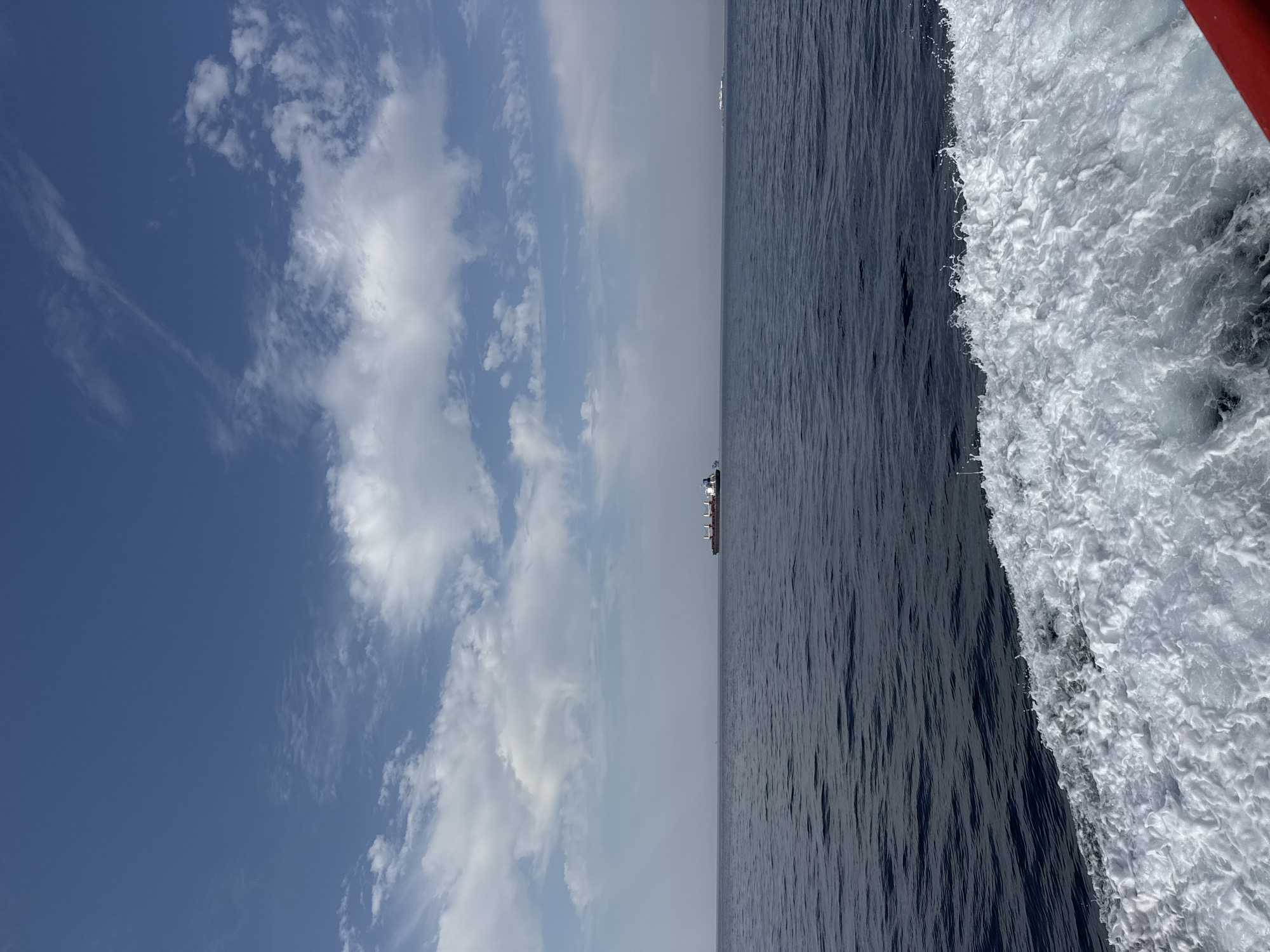 A cargo ship is visible on the horizon in the center of the frame, sailing on a dark blue sea under a partly cloudy blue sky. The foreground shows the white foam of a wake, indicating the perspective is from another moving vessel. The background is hazy, with a faint line of land visible in the distance.
