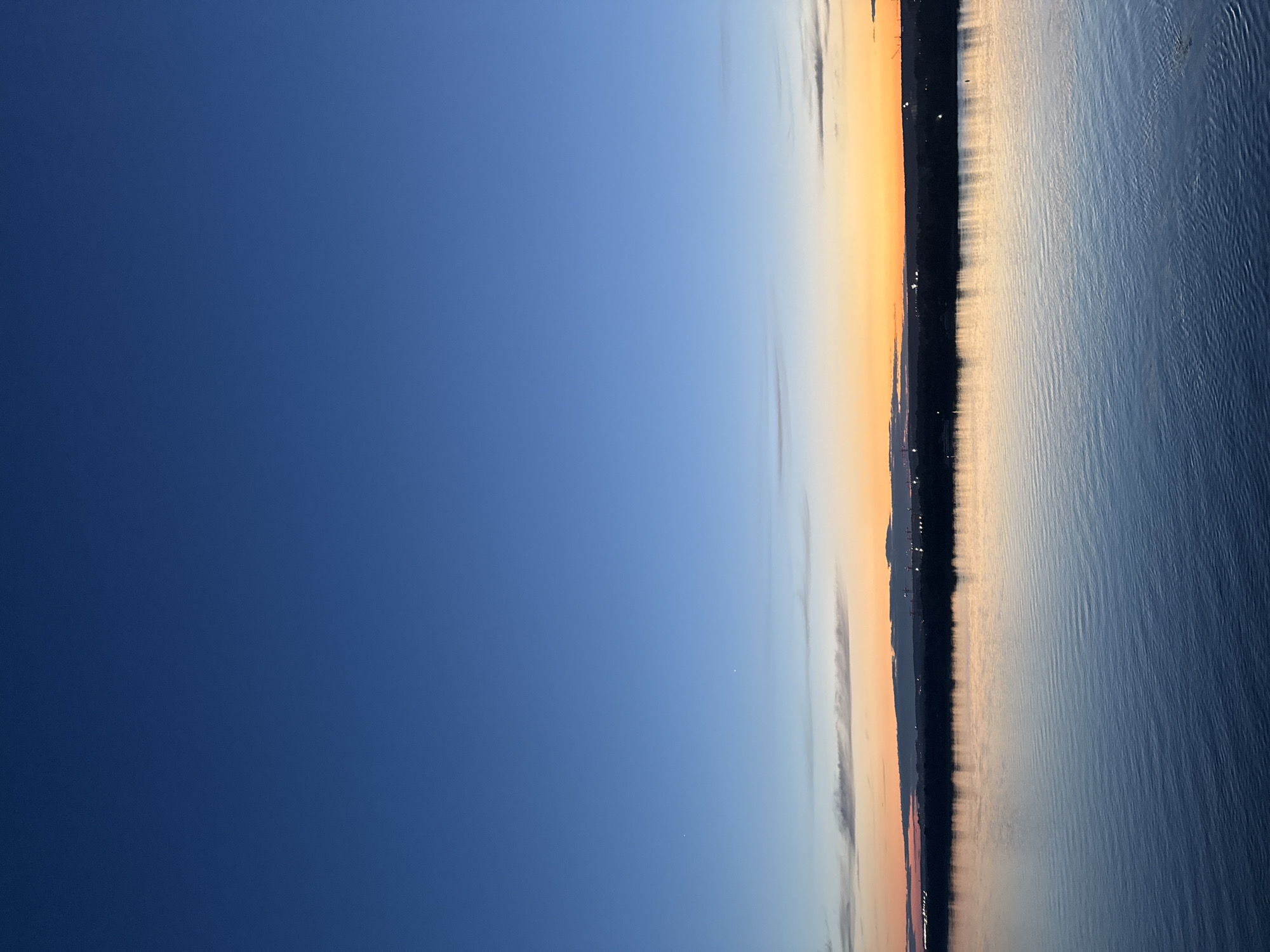 The image depicts a body of water with a dark strip of land on the horizon, suggesting a cityscape. The sky transitions from a deep blue to an orange hue, indicating either a sunrise or sunset. The water reflects the colors of the sky, showing some ripples on the surface. Based on the provided information, this image is likely taken in Perm, Russia, with the city skyline visible across the water. There are no people visible in the scene.