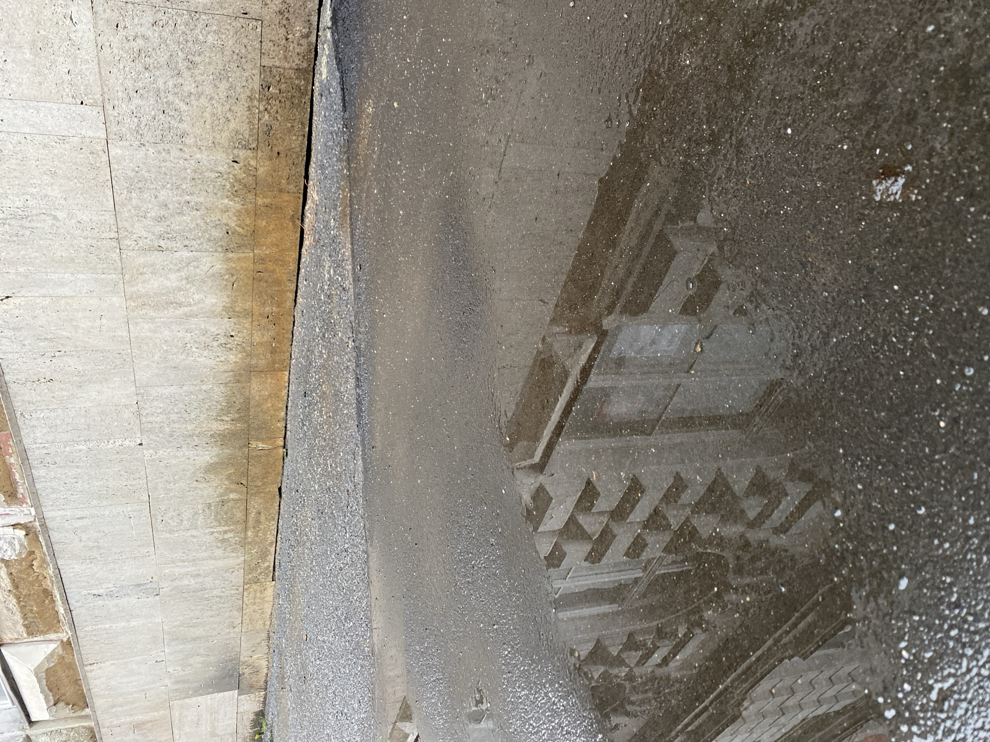 The image shows a puddle of water on asphalt next to a stone wall. The wall appears to be part of a building or a retaining wall. The asphalt is wet and reflective, showing a distorted reflection of a building with a patterned facade. The stone wall is made of large, rectangular blocks and has some staining or discoloration at the bottom. There is a strip of gravel between the asphalt and the wall. The scene is outdoors in daylight.