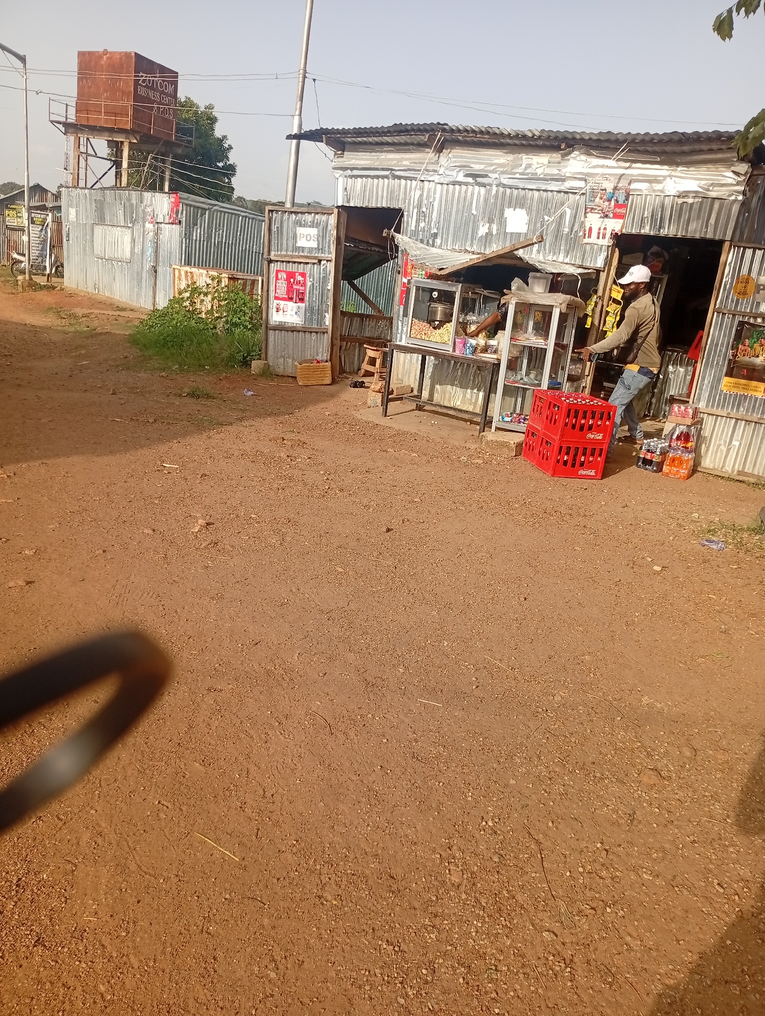 In Jos, Nigeria, a roadside shop is visible. The structure is made of corrugated metal. In front of the shop, stacked red Coca-Cola crates are prominent. A person wearing a white cap and jeans stands near the shop, seemingly interacting with a customer or attending to the business. Shelves outside the shop display various items, including beverages. To the left of the shop, a water tank tower with the inscription "ZUTCOM BUSINESS CENTER & P.O.S." stands tall. The ground is unpaved, covered with reddish-brown soil and small pebbles. Greenery is present in the background. The sky is clear and bright.