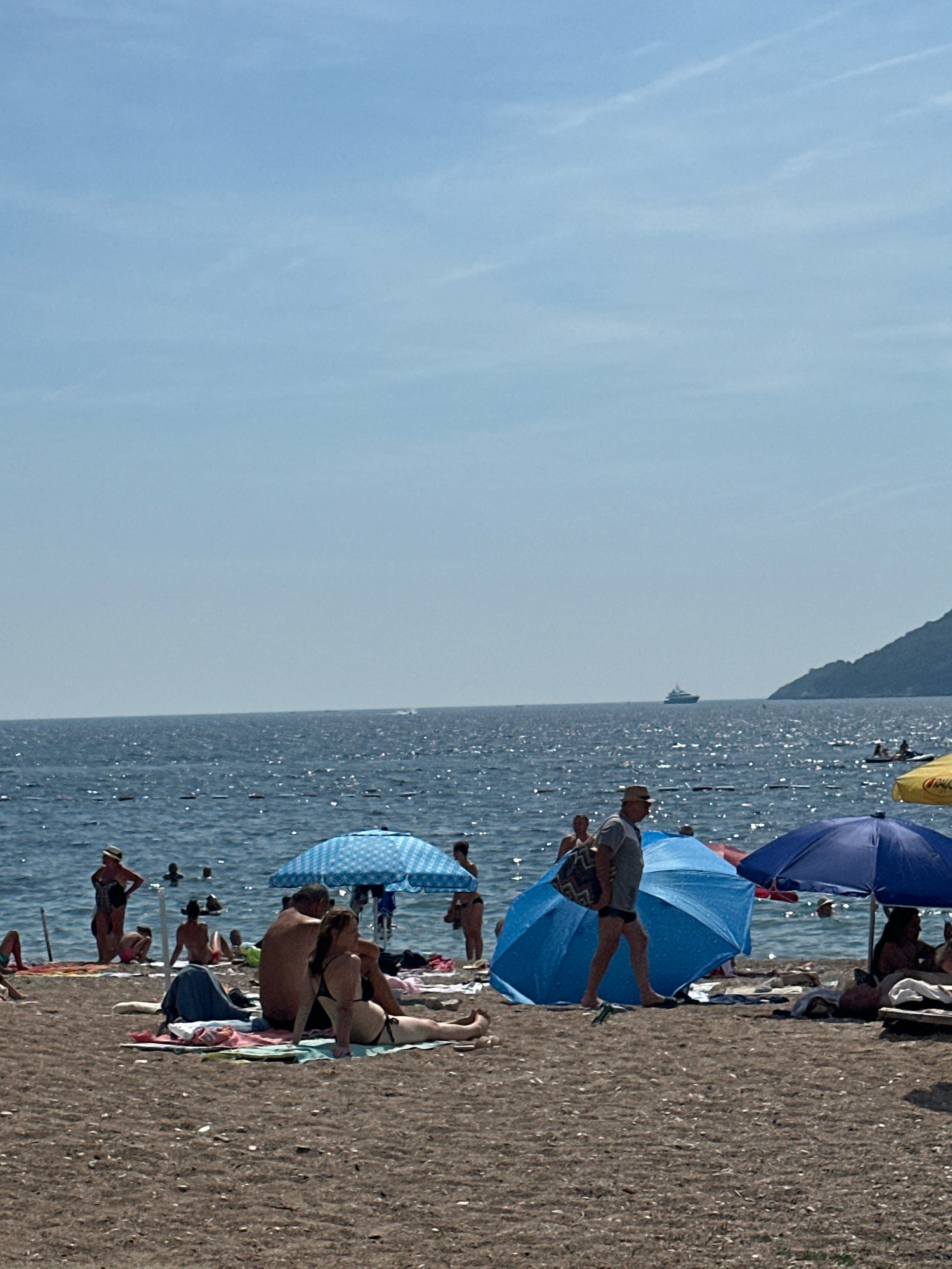 The image depicts a crowded beach scene in Budva, Montenegro, on a clear, sunny day. The calm sea stretches to the horizon, with a distant boat visible. On the sandy beach, numerous people are sunbathing, swimming, and relaxing under umbrellas. A few blue umbrellas are prominent, one with a checkered pattern. People are dressed in swimwear, and some are sitting or lying on towels. In the background, a green, hilly coastline is visible. The overall atmosphere is one of leisure and recreation on a summer day.
