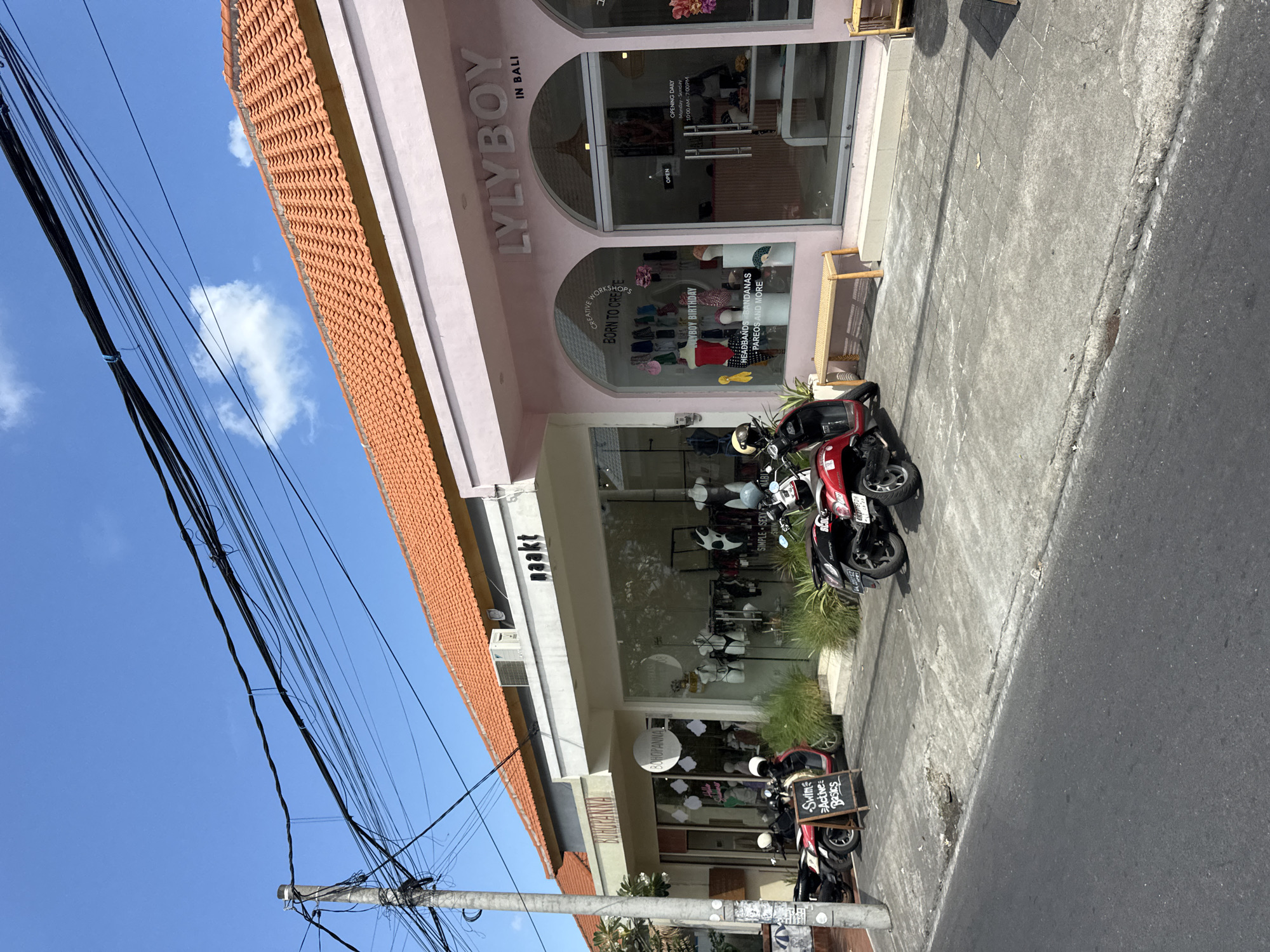 This image displays a bright, sunny street scene in North Kuta, Indonesia, featuring two commercial buildings with shops under a clear blue sky dotted with a few white clouds. Numerous black electrical wires crisscross overhead.
The prominent shop on the right, painted in a light pink color with an orange-tiled roof, is named "LYLYBOY IN BALI." Its large arched display windows showcase accessories like headbands, bandanas, and pareos, with additional text indicating "LYLYBOY BIRTHDAY" and "CREATIVE WORKSHOPS BORN TO CREATE." The shop is open daily from "10:00 AM - 7:00 PM." Inside, a display suggests "SIMPLE SEXY COOL" items. A small wooden bench is placed outside.
Adjacent to LYLYBOY is another shop with a white facade, identified by the name "naakt." A circular sign below its name also reads "BOHOPANNA." This shop's windows display clothing, and a chalkboard sign on the sidewalk advertises "Swim Active Basics."
Several red and black scooters/motorcycles are parked along the paved sidewalk in front of both stores. The scene depicts a typical commercial area during daylight hours, suggesting business operations. No people are visible in the frame.