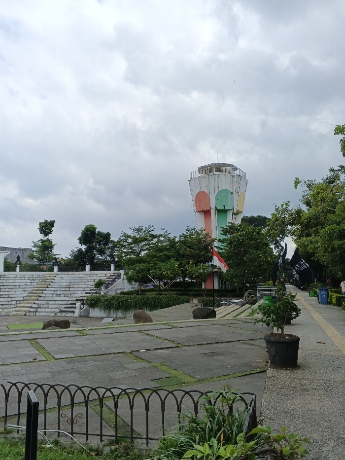 This image captures an outdoor public space in Bien Hoa, Vietnam, under an overcast sky. A tall, cylindrical tower dominates the right-center, featuring a hexagonal top and vertical panels painted in white, orange, light blue, and light green. To the left of the tower, a multi-tiered, white concrete amphitheater ascends a gentle slope. The foreground consists of a low, dark metal fence with arched sections, behind which lies a large, segmented paved area with concrete slabs separated by strips of grass. Several large, dark, smooth stones are scattered across this paved surface. A red and white flag is displayed on a pole near the base of the tower. Abundant green trees and vegetation are visible throughout the scene, notably surrounding the tower, behind the amphitheater, and lining a narrow, light-colored paved pathway on the far right. Two blue trash bins and multiple black potted plants are located along this pathway. In the upper left background, faint dark silhouettes of at least two statues are present on an elevated platform. A single small bird is visible in the upper right portion of the cloudy sky. No individuals are present in the depicted scene.