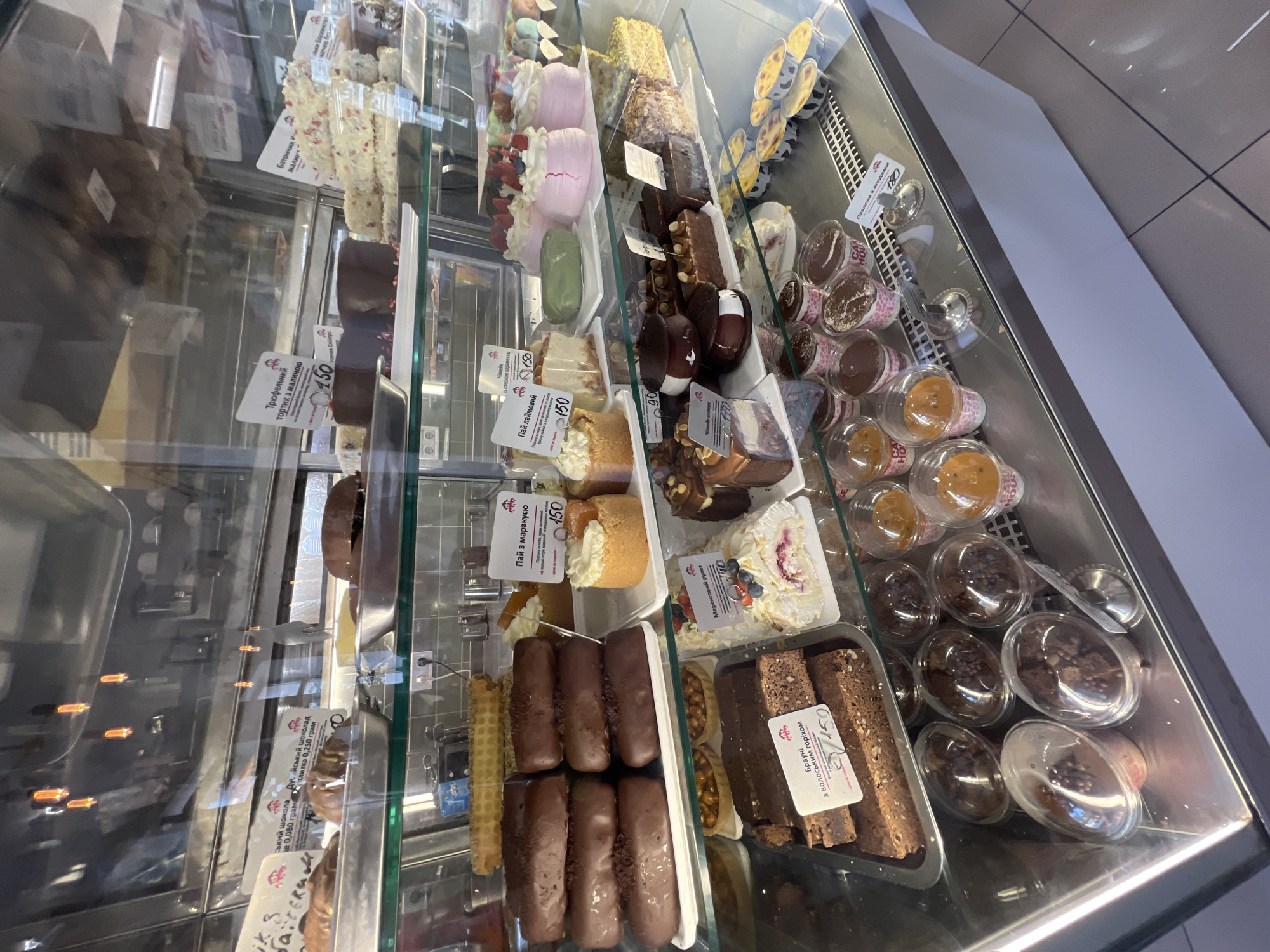 The image shows a display case filled with various pastries and desserts, likely in a bakery or cafe. The display case is made of glass and metal, and the lighting suggests an indoor environment. Several items are labelled with handwritten signs in Cyrillic script, indicating product names and prices. These include cakes, brownies, and individual dessert cups. There are no people visible in the image. The overall scene is clean and well-organized, suggesting a commercial establishment. The presence of Cyrillic script strongly suggests the location is in a Ukrainian-speaking region. Given the context of the prompt asking about Odesa, Ukraine, it is highly probable that this establishment is located in Odesa.