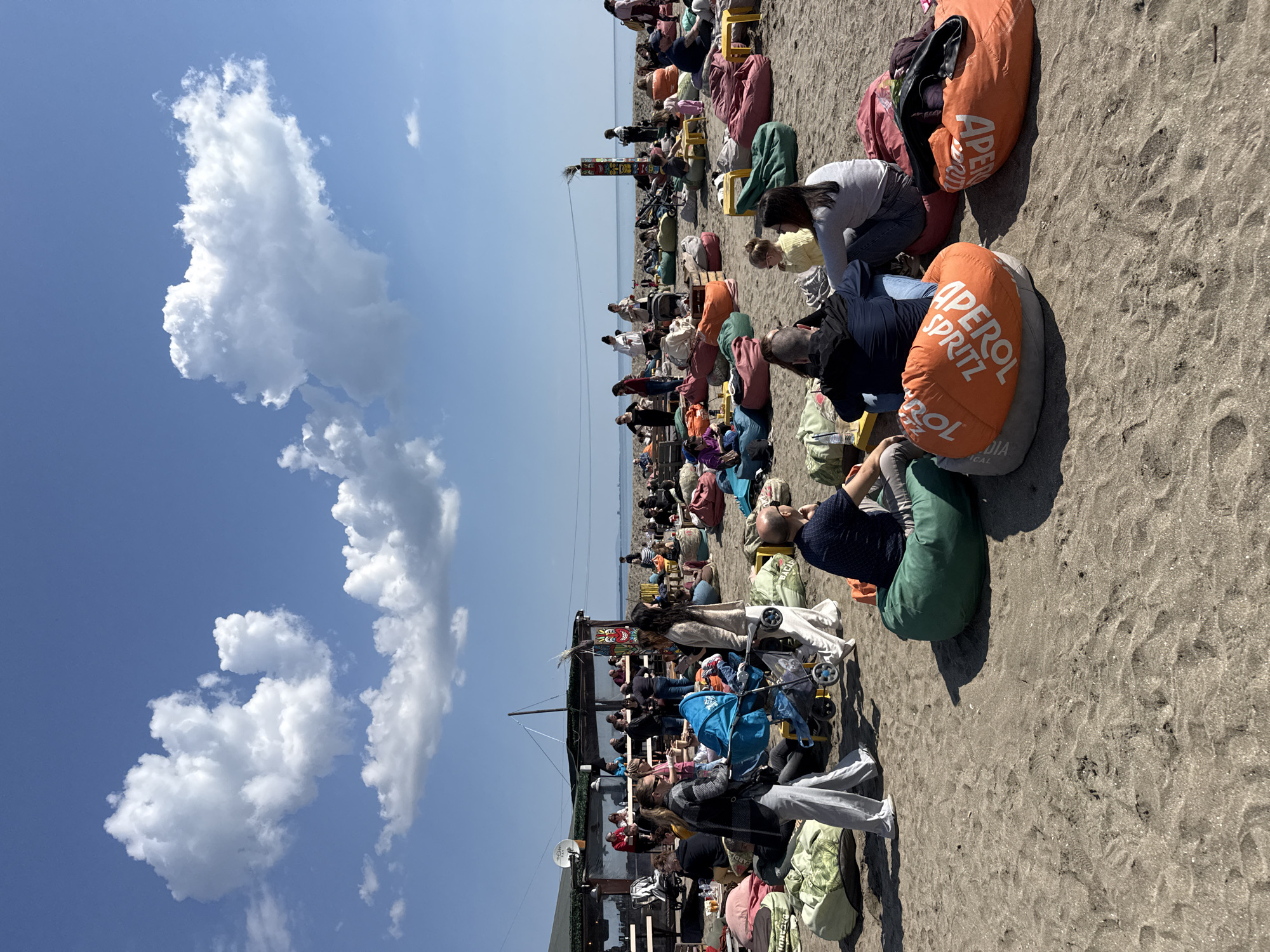 The image shows a beach scene in Burgas, Bulgaria. A large group of people are gathered on the sand, seated on colorful beanbag chairs, many of which are branded "Aperol Spritz." The crowd extends towards a structure that appears to be a beach bar or cafe. Some individuals are interacting with others nearby, and some are using small, yellow stools. In the background, a body of water stretches to the horizon under a light blue sky. To the left, cumulus clouds are visible. The beach itself has a coarse, sandy texture. A few people are walking near the bar, and a baby stroller is among the objects.