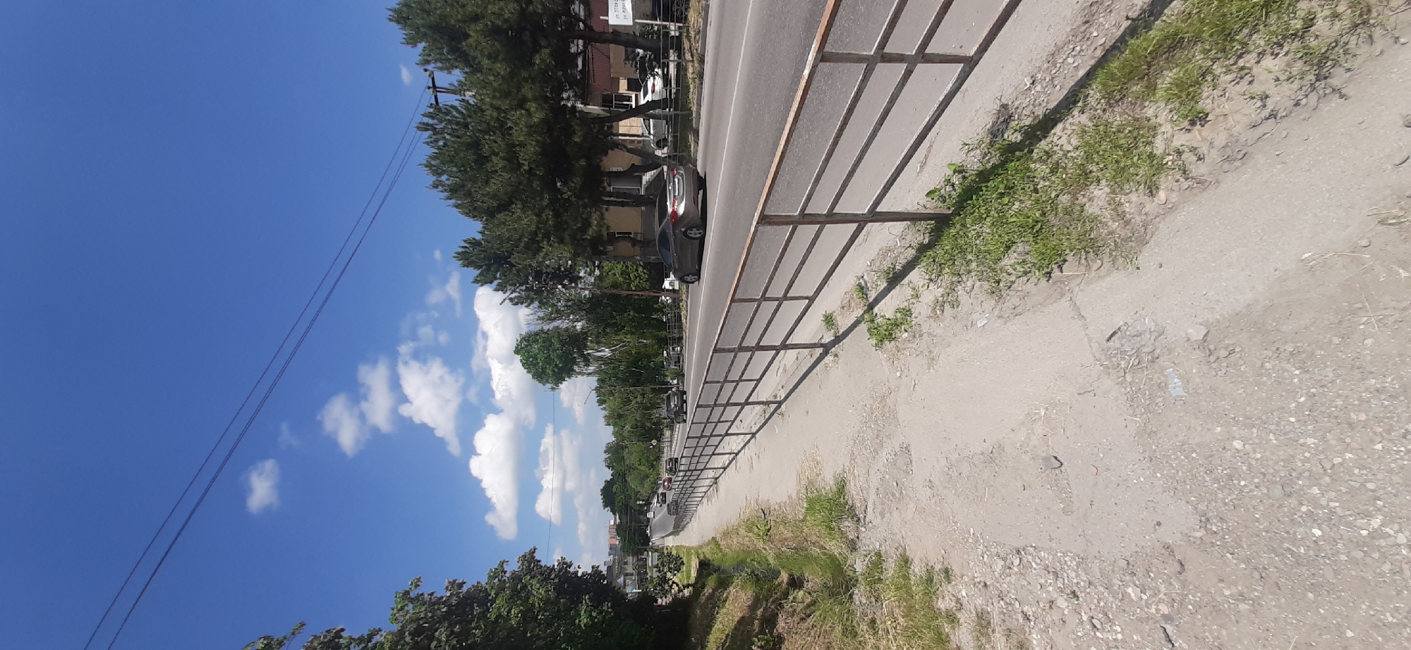 The image shows a street scene in Yessentukskaya, Russia. A paved sidewalk runs along the left side of the frame, bordered by a metal railing and a grassy ditch. Beyond the railing is a road with several cars driving in the same direction. Trees line the right side of the road, with buildings visible behind them. The sky is blue with scattered white clouds, and power lines are visible overhead. The lighting suggests it is daytime, possibly late morning or early afternoon. The overall impression is of an ordinary urban street on a clear day.