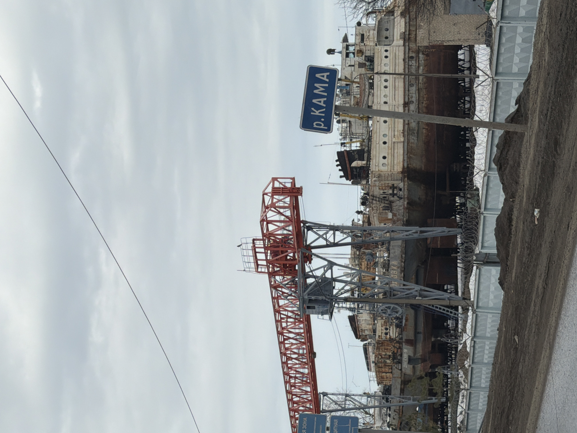 The image shows a red and grey overhead crane in the city of Chaikovsky, Russia. A sign labeled "p. KAMA" (River Kama) is visible, confirming the location. In the background are several derelict, rusted boats or ships, enclosed by a fence with barbed wire. A power line stretches across the upper left part of the image. The sky is overcast. A road or walkway is visible in the lower right of the image.