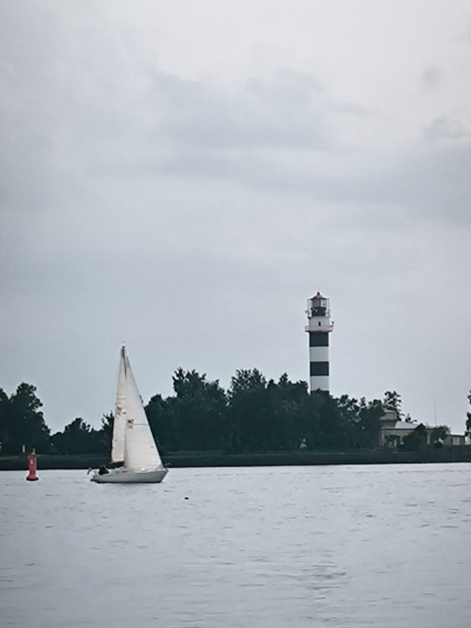 A sailboat is depicted on a body of water, with a lighthouse and trees on the shore in the background. The sky is overcast. The lighthouse has black and white stripes. The sailboat has a white sail. A red buoy is visible in the water to the left of the sailboat. The scene is likely Riga, Latvia, given the presence of the Daugava River and the proximity of the Daugava Lighthouse.