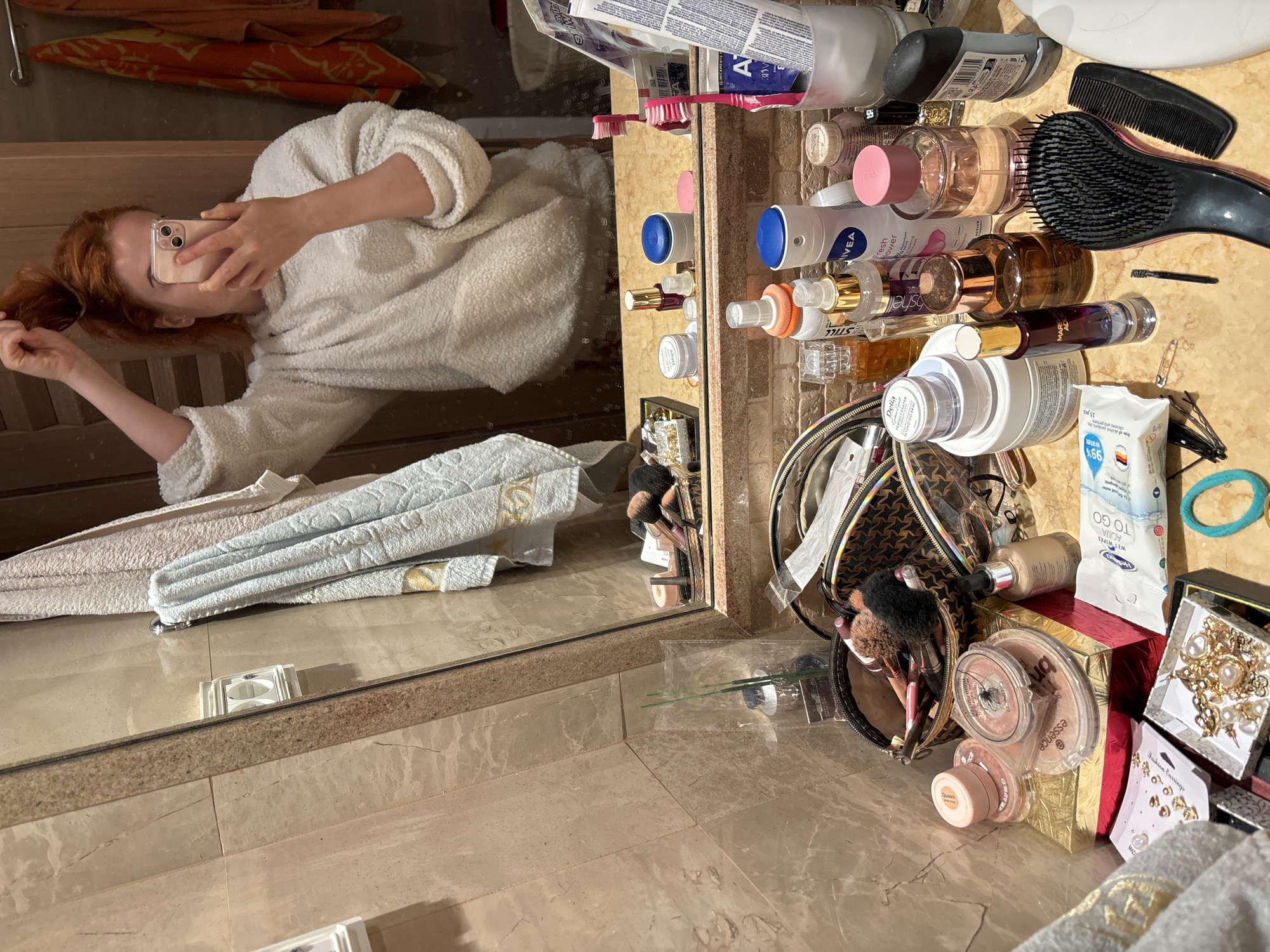 The image shows a woman taking a selfie in front of a mirror in what appears to be a bathroom. The woman has red hair and is wearing a white robe. In the mirror, we can see her holding a phone up to take the picture.
The bathroom countertop is cluttered with various items. These include toiletries such as toothpaste, toothbrushes, lotion bottles, and perfumes. There are also hair care items like combs and hair accessories. Makeup products are visible as well, including brushes, foundation, blush, and eyeshadow. A makeup bag is also present. Additionally, there are jewelry boxes containing earrings and necklaces. The countertop and floor have a marble-like finish.
Based on the available information, it's impossible to confirm that the scene is located in the city of Budva, Montenegro.
