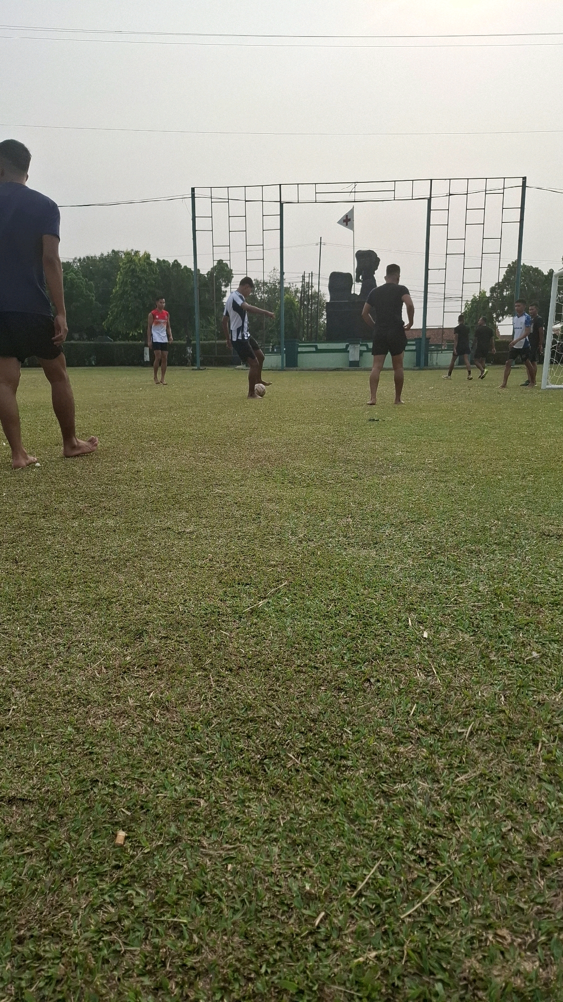 This is a full, eye-level shot of a group of men playing soccer on a grassy field. The field is mostly green with some patches of brown. In the background, there is a white fence with a net and a goalpost. There is also a large, dark sculpture in the background, behind the fence, with a white flag on top of it. The sky is overcast. One man in the foreground, on the left, is wearing a dark blue shirt and black shorts and is barefoot. He is facing away from the camera. Another man in the middle of the frame is wearing a white shirt with black stripes and black shorts and is kicking a soccer ball. He is also barefoot. To the right of him, another man is wearing black shorts and a black shirt. Several other men in similar attire are scattered across the field, some of them are partially obscured by the goalposts. The location is identified as Sukaraja, Indonesia.