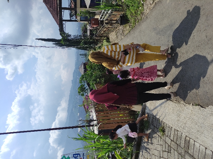 The image depicts a group of four people walking on a paved path in a rural area, located in Kecamatan Pujon, Indonesia, at the coordinates -7.854 latitude and 112.454 longitude. Three of the people are women, wearing headscarves. Two women are walking in the foreground, side-by-side. The woman on the left is wearing a burgundy headscarf and a maroon cardigan over a white shirt and dark pants. The woman on the right is wearing a gold headscarf, a yellow and white striped top, and yellow pants. A girl in a pink and white dress is walking behind the two women. There is a younger girl with short dark hair, wearing a white shirt, walking in the opposite direction, in the background of the image. There is a light-colored wooden fence on the left side of the image. In the background, there are green trees, houses, and a blue sky with white clouds. The sun is shining. A gray trash can is visible in the lower right corner of the image, on a grassy patch adjacent to the paved path.