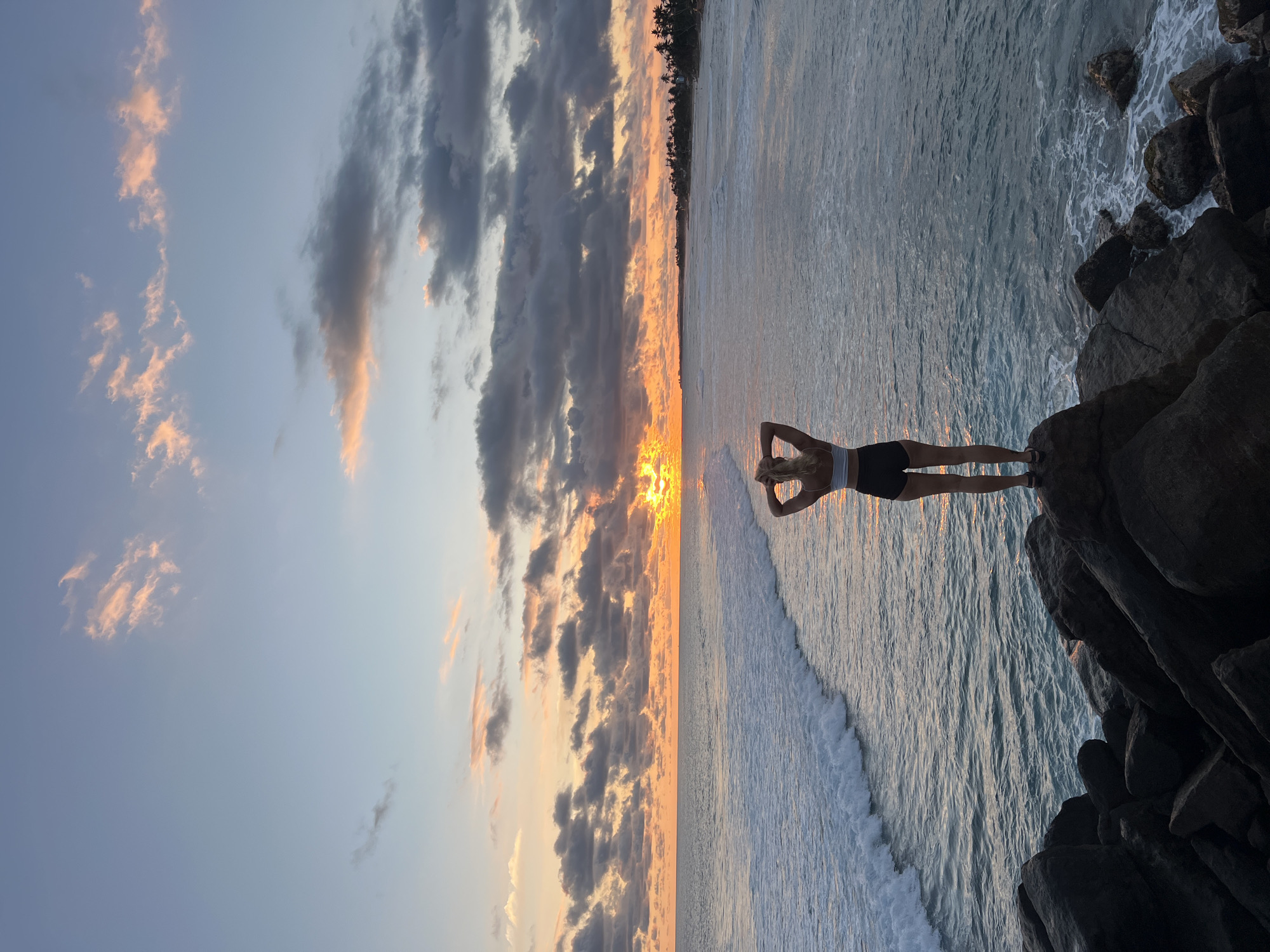 A woman stands on rocks overlooking the ocean, silhouetted against a sunset at Goviyapana, Sri Lanka. The sky has a mix of light blue with light orange clouds and dark storm clouds. The sun is setting on the horizon. The ocean has a light blue tint with white waves. The woman is wearing a white top and black shorts with her hands behind her head. She is standing on dark rocks.