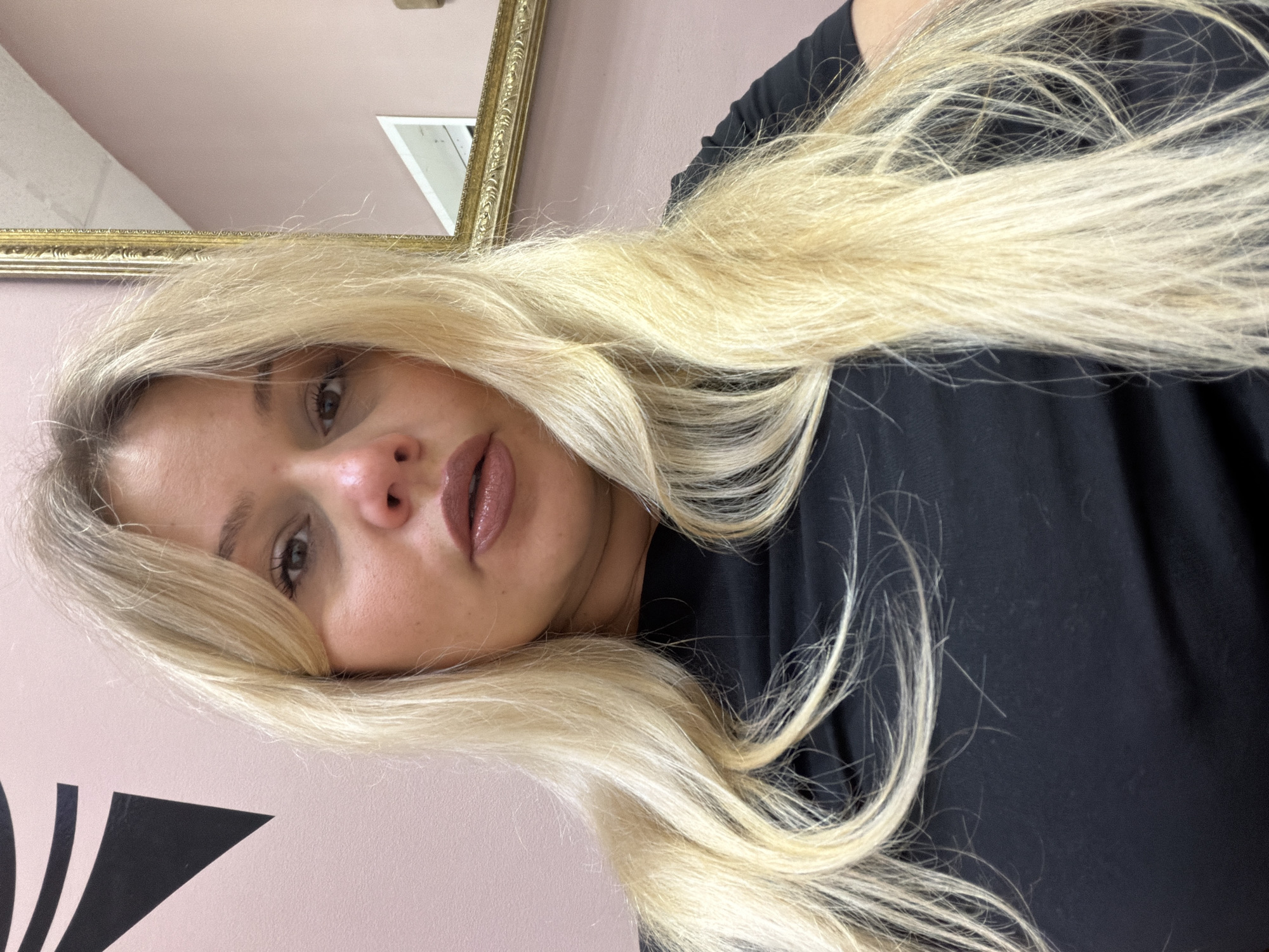 A woman with long, blonde hair is shown close-up, wearing a black shirt. The background includes a light pink wall, possibly with black decorative elements, and a gold-framed mirror partially visible. The location is not discernible from the image.