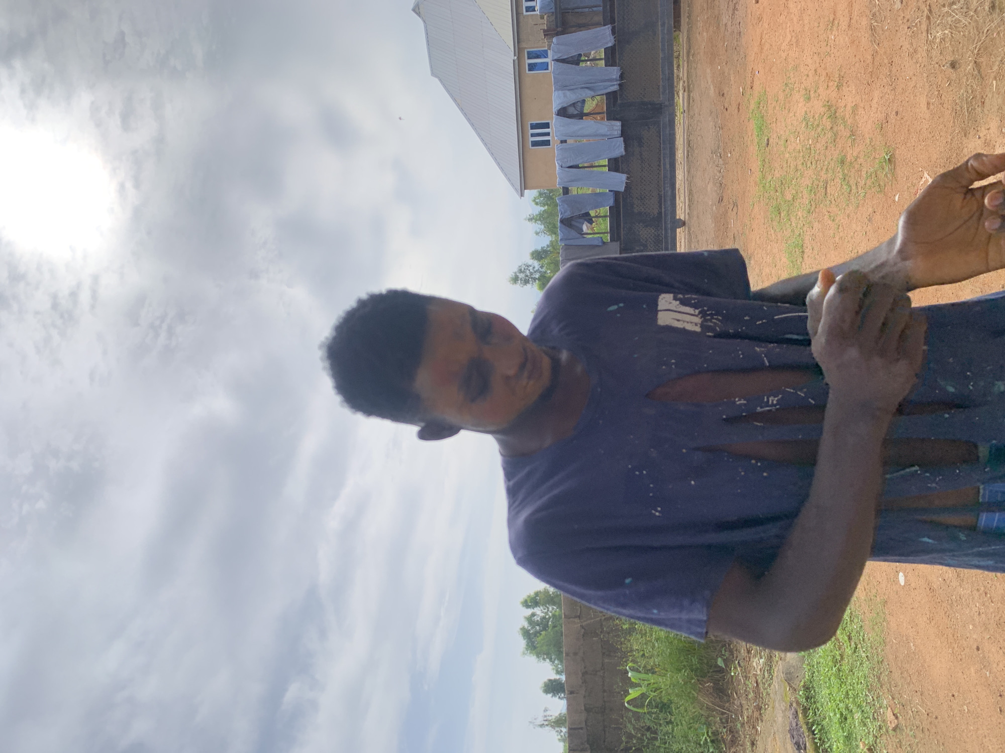 An outdoor, medium shot captures a young Black male standing in what appears to be a rural or semi-urban area in Gwafan, Nigeria. The subject is looking down at his left wrist and arm, which he is touching with his right hand. He is wearing a dark blue t-shirt with several large tears, revealing his torso. The shirt is also splattered with white and teal paint. His hair is short and dark. The background shows a dirt ground, a low brick wall with green foliage behind it, and a metal gate with several pairs of light blue pants hanging on it. Beyond the gate, a building with a light brown facade and a silver roof is visible. The sky is cloudy, with some sunlight filtering through.