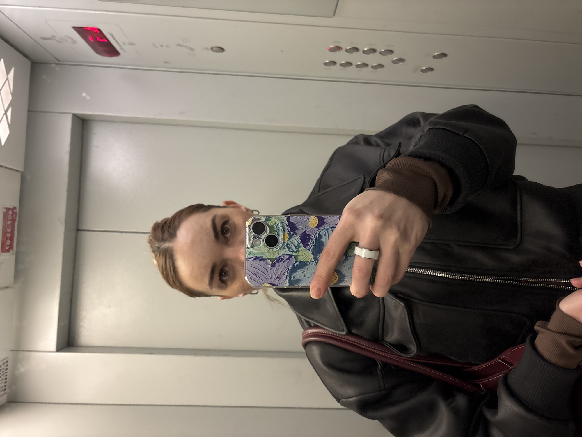 A person takes a selfie in what appears to be an elevator. The elevator has white panels, a floor indicator showing the digit "1", and a panel of buttons. The person is wearing a black leather jacket over a brown long-sleeved shirt and has a ring on their finger. They are holding a smartphone with a decorative case in front of their face. A glimpse of a maroon-colored bag strap is also visible. The image does not provide information about the city of Saint Petersburg, Russia.