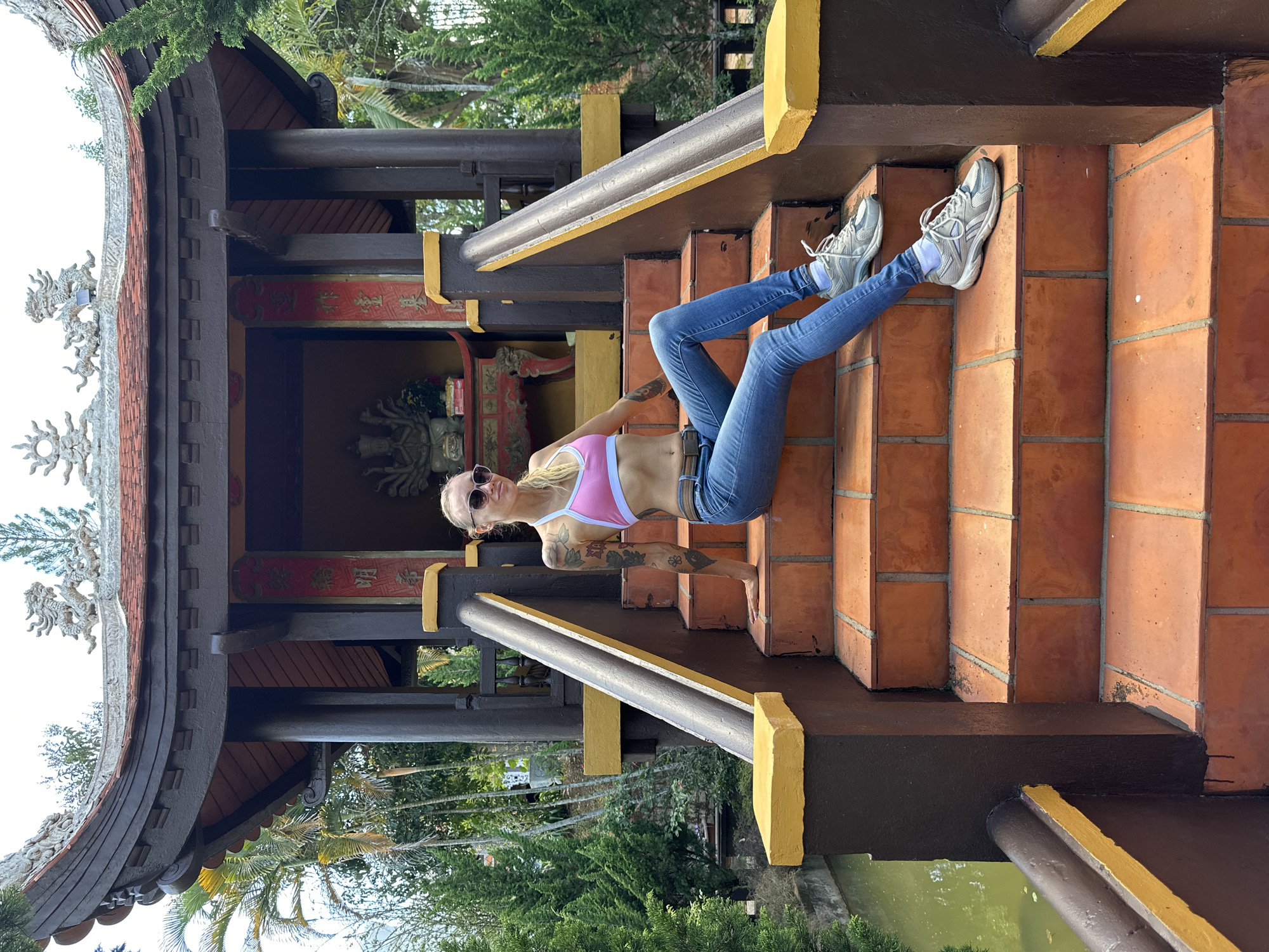 This media file features a young woman with blonde hair and sunglasses, wearing a pink sports bra and blue jeans, sitting on a set of red brick stairs. She has tattoos on her arms. The stairs are part of an outdoor structure with a dark brown and yellow color scheme, reminiscent of East Asian architecture, featuring decorative elements like dragon carvings. Behind her, a glimpse of a statue can be seen within an alcove adorned with red banners featuring Vietnamese text. The setting appears to be a temple or a culturally significant site in Vietnam, possibly Thôn 5. Lush green trees and foliage surround the structure, with a body of water visible in the lower portion of the image. The lighting suggests it is daytime, with sunlight casting shadows on the stairs, indicating fair weather. The woman's pose and expression convey a relaxed, casual demeanor.