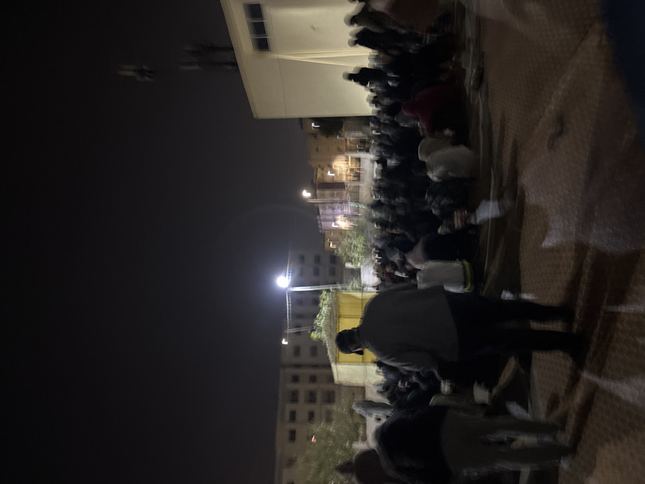 This image depicts a nighttime outdoor gathering in an urban area of Fez, Morocco. Numerous individuals, estimated to be several dozen, are assembled on a patterned paved surface. Most are seated on the ground, while a few figures are standing. A male subject in a light grey jacket and dark cap is prominently featured in the foreground, facing away from the camera. Another person is partially visible in the left foreground, also standing.
In the mid-ground, a temporary structure, possibly a tent or canopy, with yellow fabric is visible. The scene is illuminated by multiple bright streetlights, one centrally prominent on the left, which cast elongated shadows. The background features several multi-story buildings, likely residential, with some illuminated windows. To the far right, a distinct lighter-colored building is visible, topped with a cellular antenna array or similar communication equipment. The overall image exhibits significant motion blur, suggesting movement of subjects or a low-light photographic condition. The sky above is dark and cloudless.