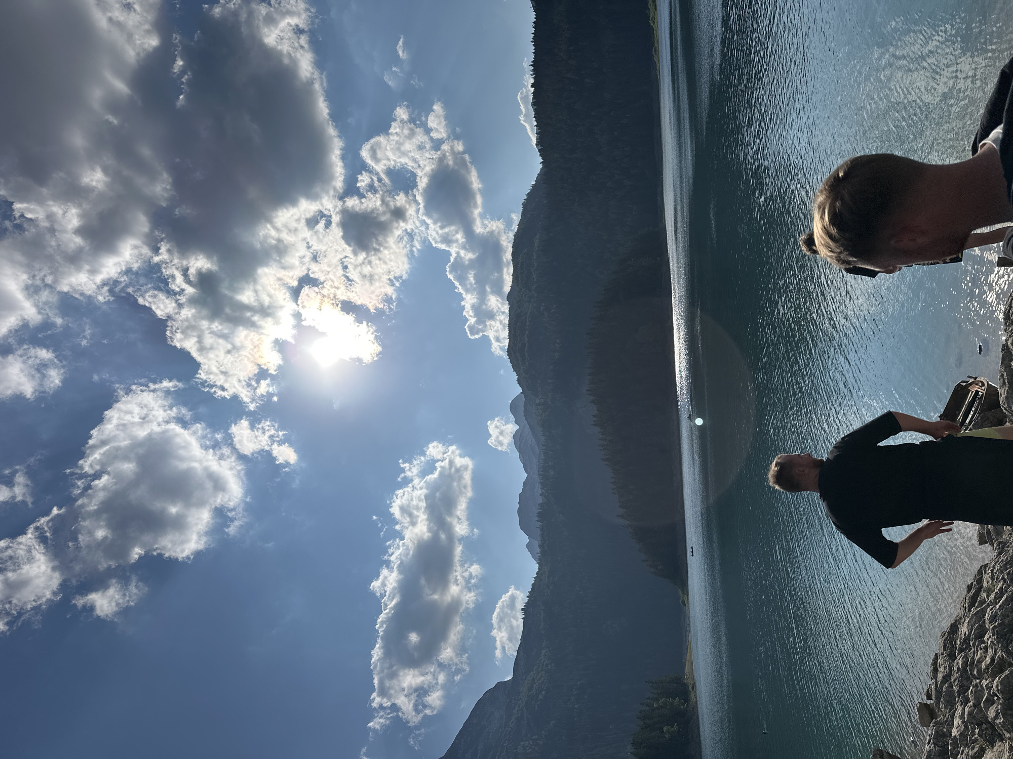 The image depicts a scenic mountain lake environment. In the foreground, two individuals are visible. The person on the left, facing away from the camera, is wearing a black t-shirt and has their hands on their hips. They are standing on a rocky shore. To the right of this individual, another person's head and shoulders are visible, also facing away from the camera, with light-colored hair. The lake occupies the midground, appearing calm with a shimmering surface reflecting the sunlight. In the background, a dense forest covers the slopes of majestic mountains, with several peaks silhouetted against a bright, partly cloudy sky. The sun is positioned high in the sky, partially obscured by clouds, casting a bright light and creating glare on the water. The overall impression is one of a tranquil, natural landscape, likely situated in a mountainous region. The location is identified as Pitomine, Montenegro.