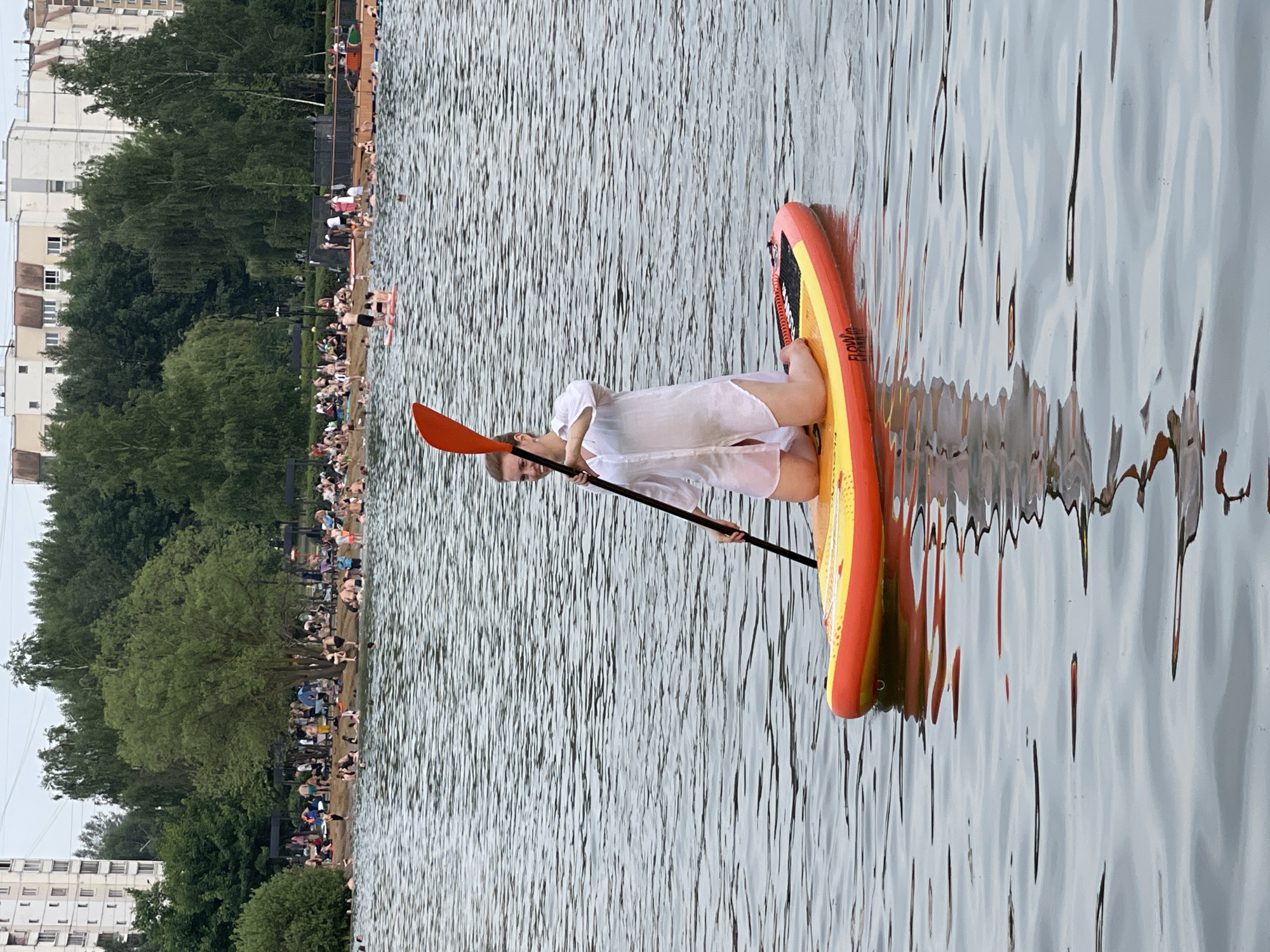 A woman kneels on a yellow and orange paddleboard in the foreground of a lake with rippling grey water. She is wearing a white, sheer button-down shirt and dark shorts. She holds a black paddle with an orange blade, positioned in front of her. In the background, a crowded beach and park area are visible, with many people sitting and standing on a sandy shore and swimming in the water. Tall, green trees line the background, with buildings of varying heights visible behind them. The overall scene appears to be a public park and lake within the city of Moscow, Russia, likely during a warm weather season.