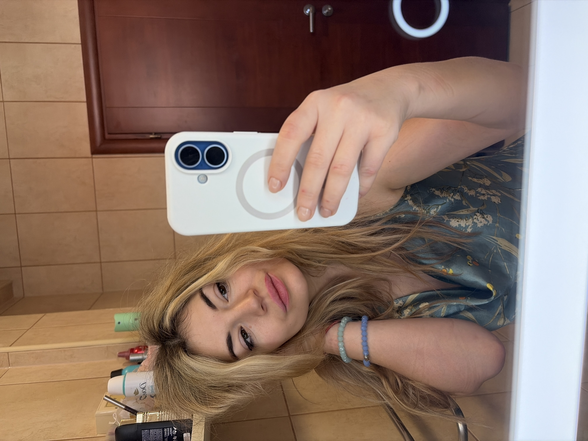 A woman is taking a selfie in a bathroom. She holds a white cell phone with two cameras and a magnetic circle in her right hand. The woman has long blonde hair, is wearing two beaded bracelets on her left wrist, and appears to be wearing a teal floral print top. The bathroom has tan tiled walls, a dark wood cabinet, and a white ring light is visible above the cabinet. A shelf with toiletries is to the left of the woman. The precise location in Budva, Montenegro cannot be determined from the image.