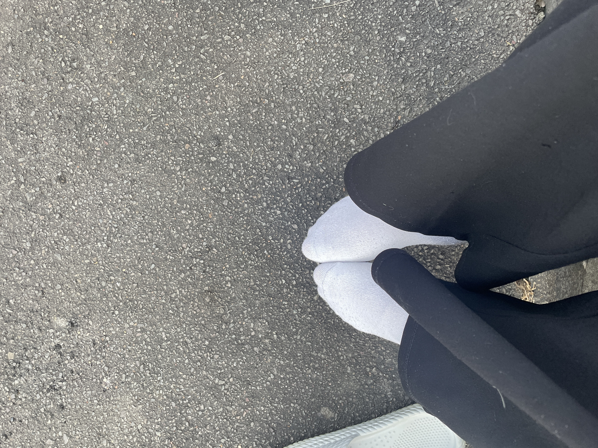 The image shows a close-up, overhead view of a person's legs and feet standing on asphalt. The person is wearing white socks and black pants. To the left of the frame, a portion of a white shoe or sandal is visible. The asphalt appears to be a typical road surface, with small stones and debris. There is no indication of the location in Wiehl, Germany within the image itself. The scene is static and focused on the lower extremities of an individual on an outdoor surface.