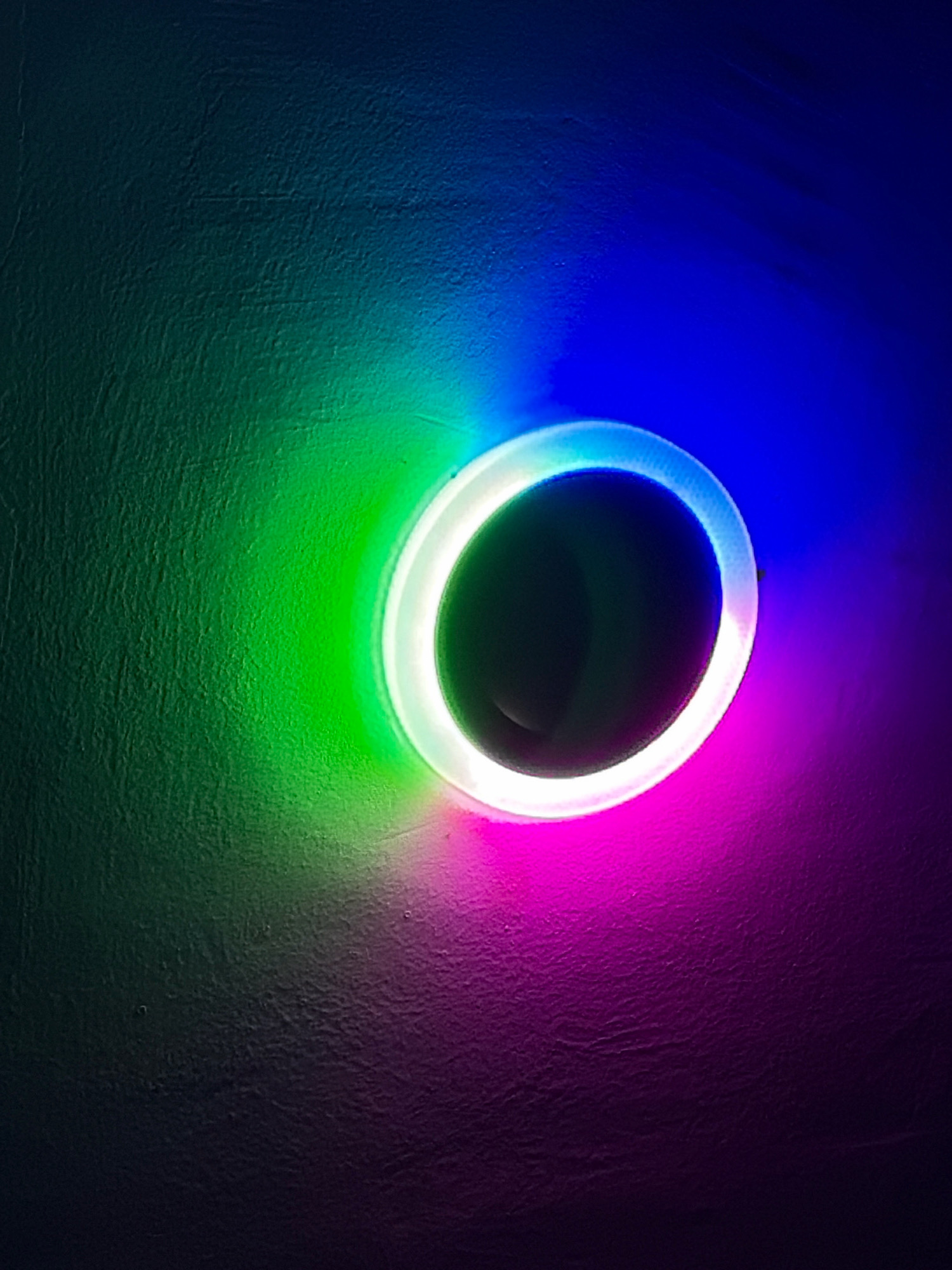The image shows a circular light fixture with a white outer ring emitting green, blue, and pink light onto a textured, dark surface, likely a wall. The lighting is abstract and does not depict any specific location in Bodma, India.