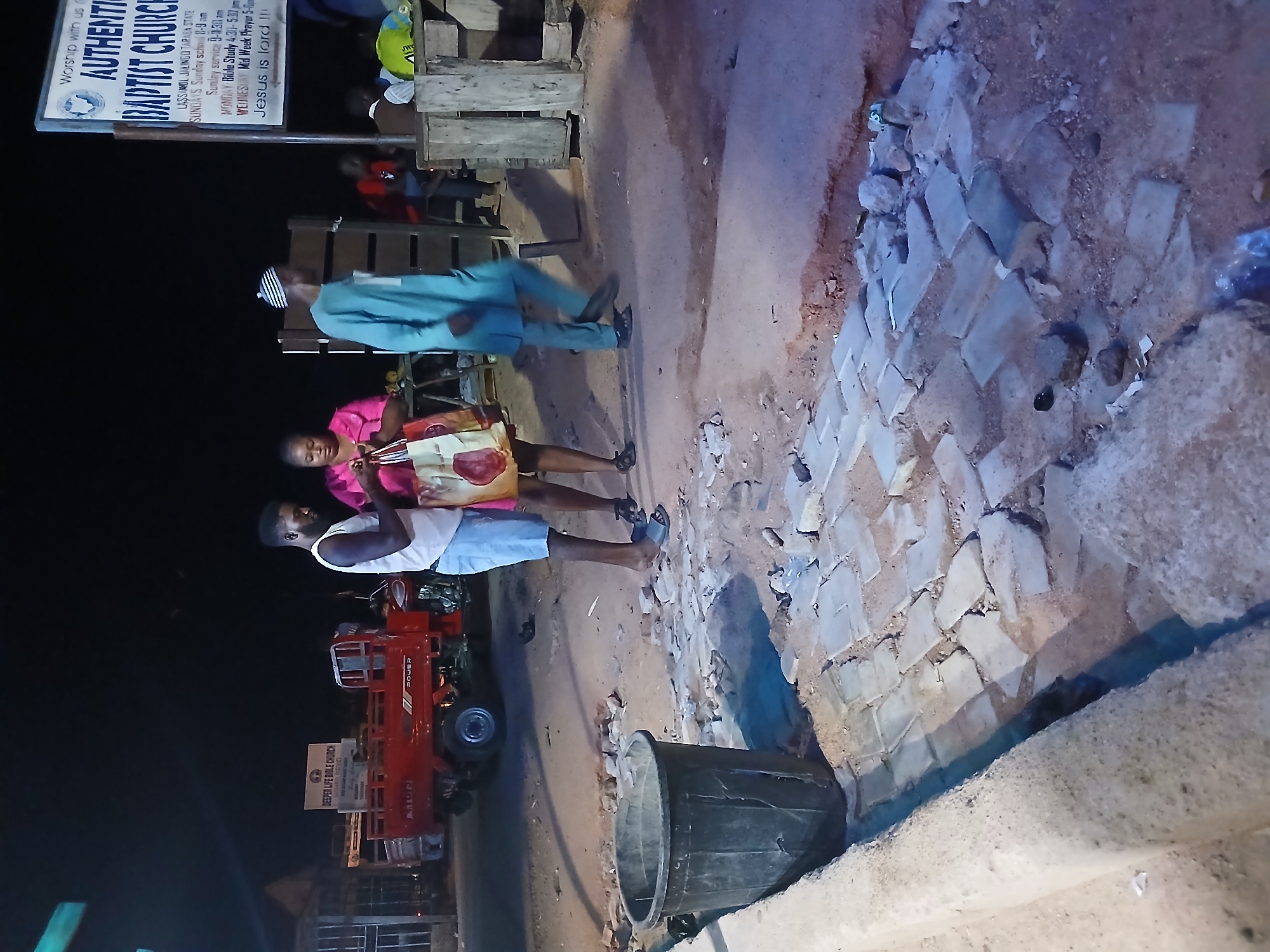 The image depicts a street scene at night in Jalingo, Nigeria, in front of the Authentic Baptist Church. A red three-wheeled vehicle is parked on the street. In the foreground, a large black trash bin sits next to a sidewalk, with broken paving stones scattered on the ground, indicating ongoing or recently completed construction or repair work. Three people are visible: a man and a woman standing together, with the woman holding a patterned shopping bag, and a man in a blue traditional outfit walking away from the camera. Further in the background, more people are gathered near the church entrance and a wooden structure. A sign for "Deeper Life Bible Church" is also visible in the background, to the left of the main church sign. The environment appears to be a dusty, unpaved area alongside a paved section of road.