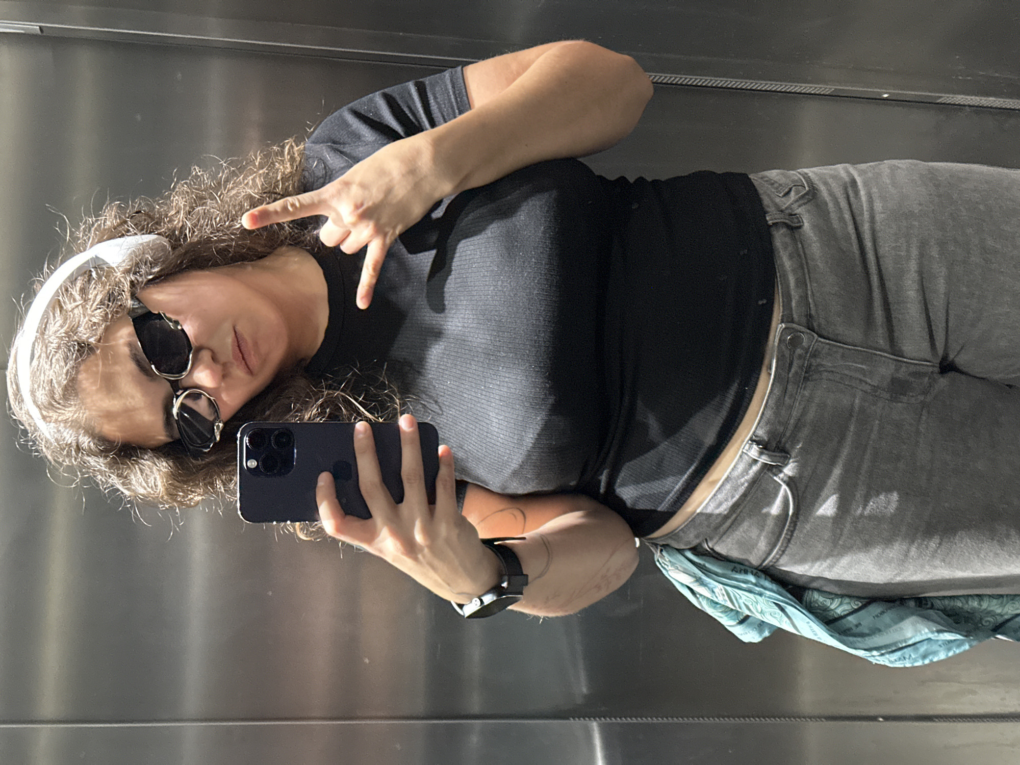 A woman takes a selfie in an elevator. She wears a black short-sleeved shirt, gray jeans, a white headband, and sunglasses. Her left hand holds a black smartphone. Her right hand forms a peace sign. A blue patterned scarf hangs from her jeans. She wears a wristwatch on her left wrist. The elevator interior is metallic. The location is unknown, and there is no information about it being in Budva, Montenegro.