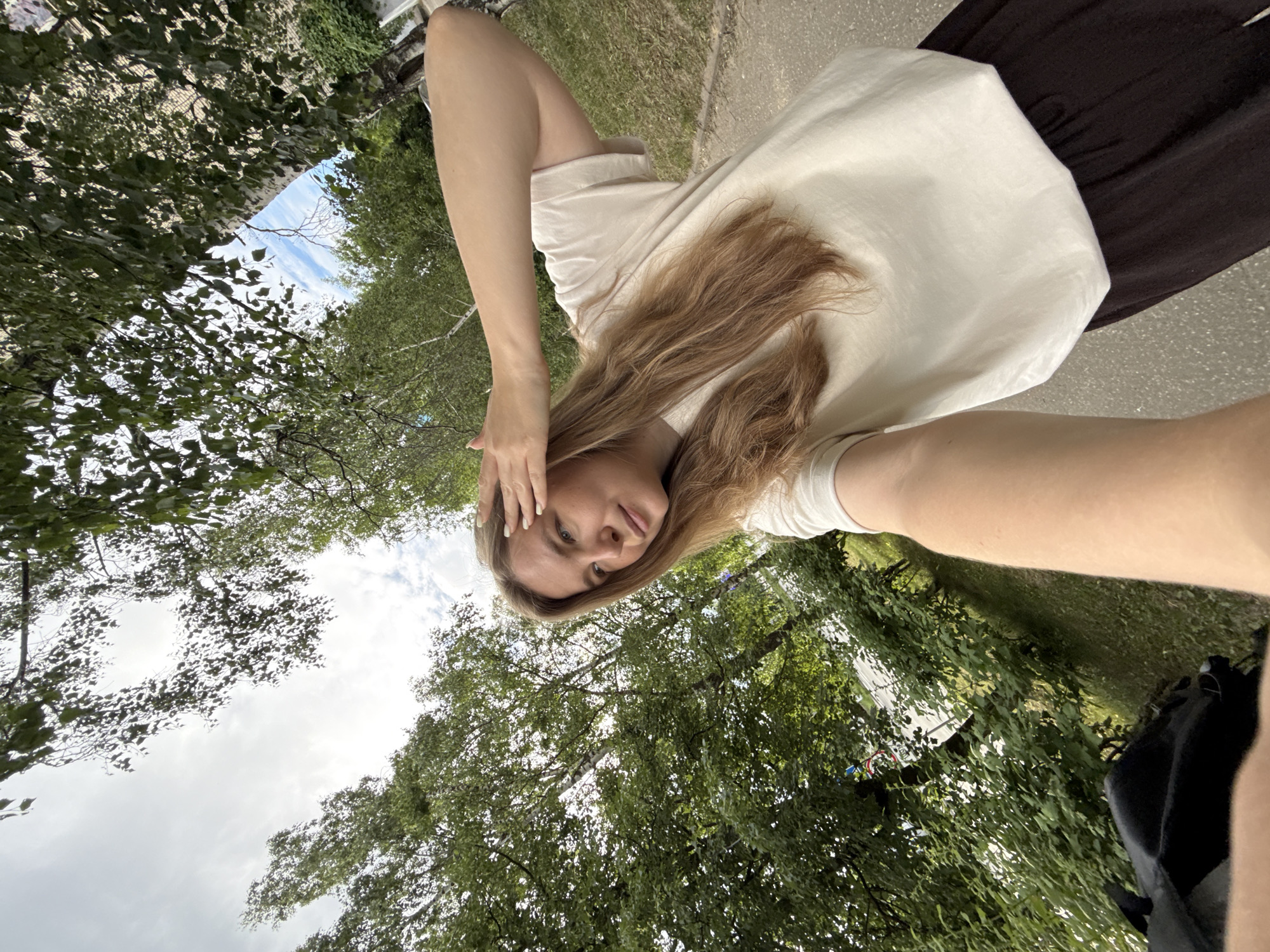 A young woman with long, light brown hair is taking a selfie outdoors. She is wearing a light-colored t-shirt and dark pants. Her right hand is raised to her forehead, and she is looking off to the side. The background consists of lush green trees and a cloudy sky. A paved path is visible to her right, and a black bag is partially visible in the bottom left corner. The scene appears to be a park or a street lined with trees.