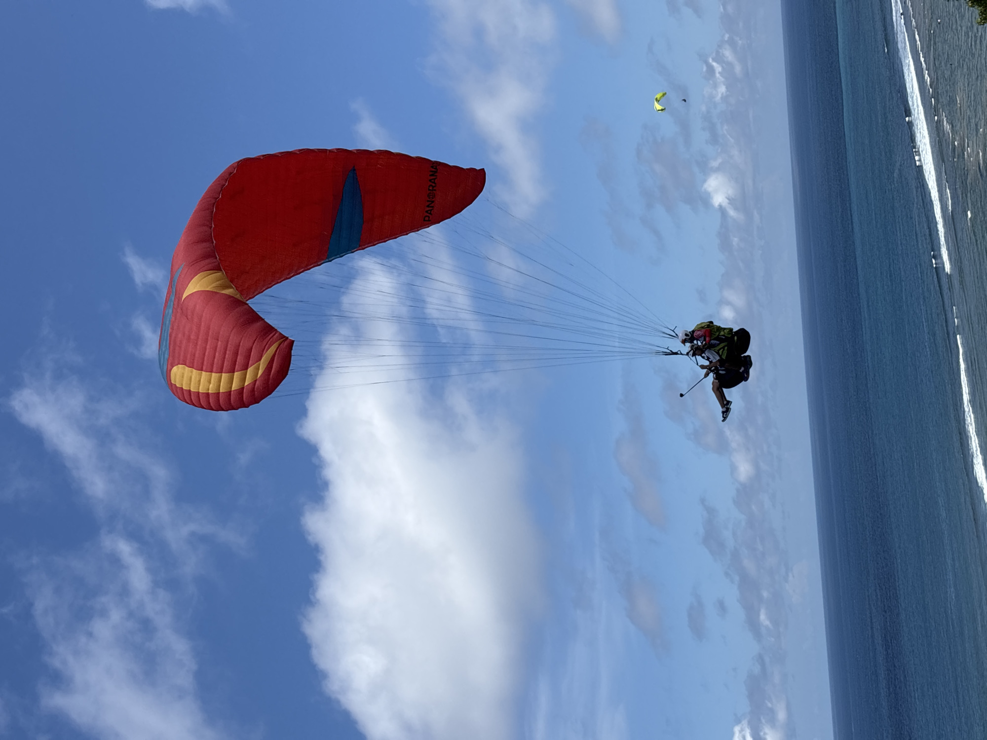 A person is paragliding over the water near the city of Benoa, Indonesia. The paraglider has a red, yellow, and blue canopy. The person is wearing a black harness and black shoes. The sky is blue with white clouds. The water is blue. There is another yellow paraglider in the distance. Some trees are seen to the right of the image.