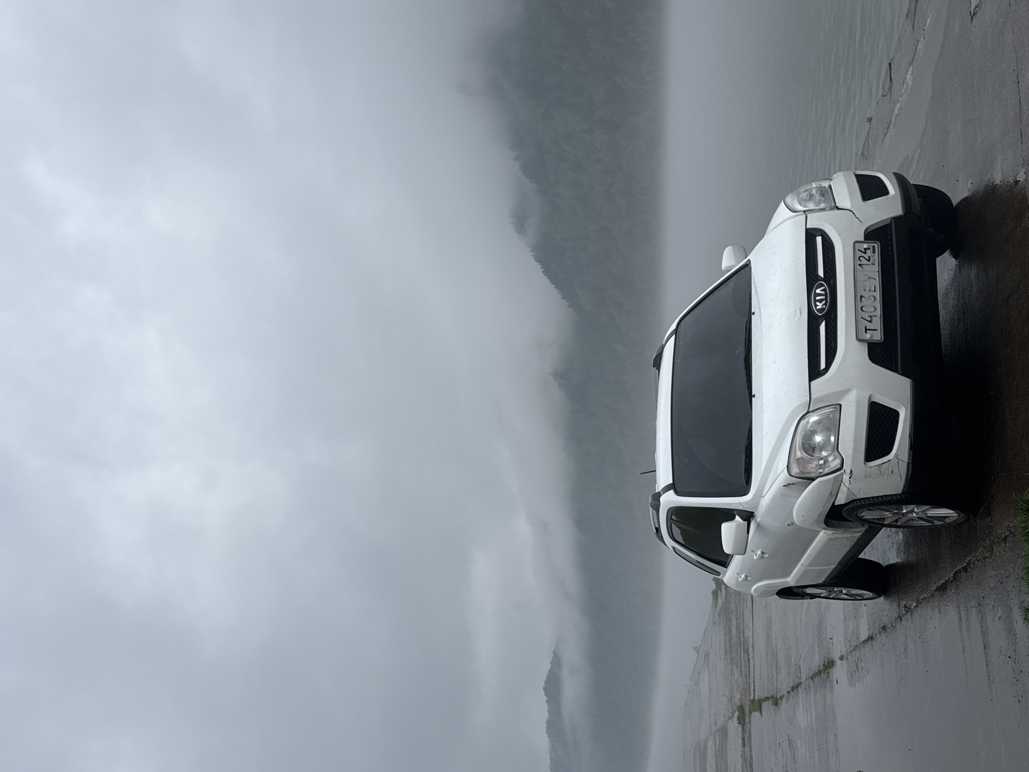 A white Kia Sportage is parked on a wet concrete surface adjacent to a body of water, possibly the Yenisey River. The environment is overcast and foggy, obscuring the view of the surrounding landscape, which includes distant mountains. The location is likely in or near Divnogorsk, Russia, as indicated by the license plate "T403EY 124" on the vehicle, where 124 is the Russian code for the Krasnoyarsk region. No people are visible.