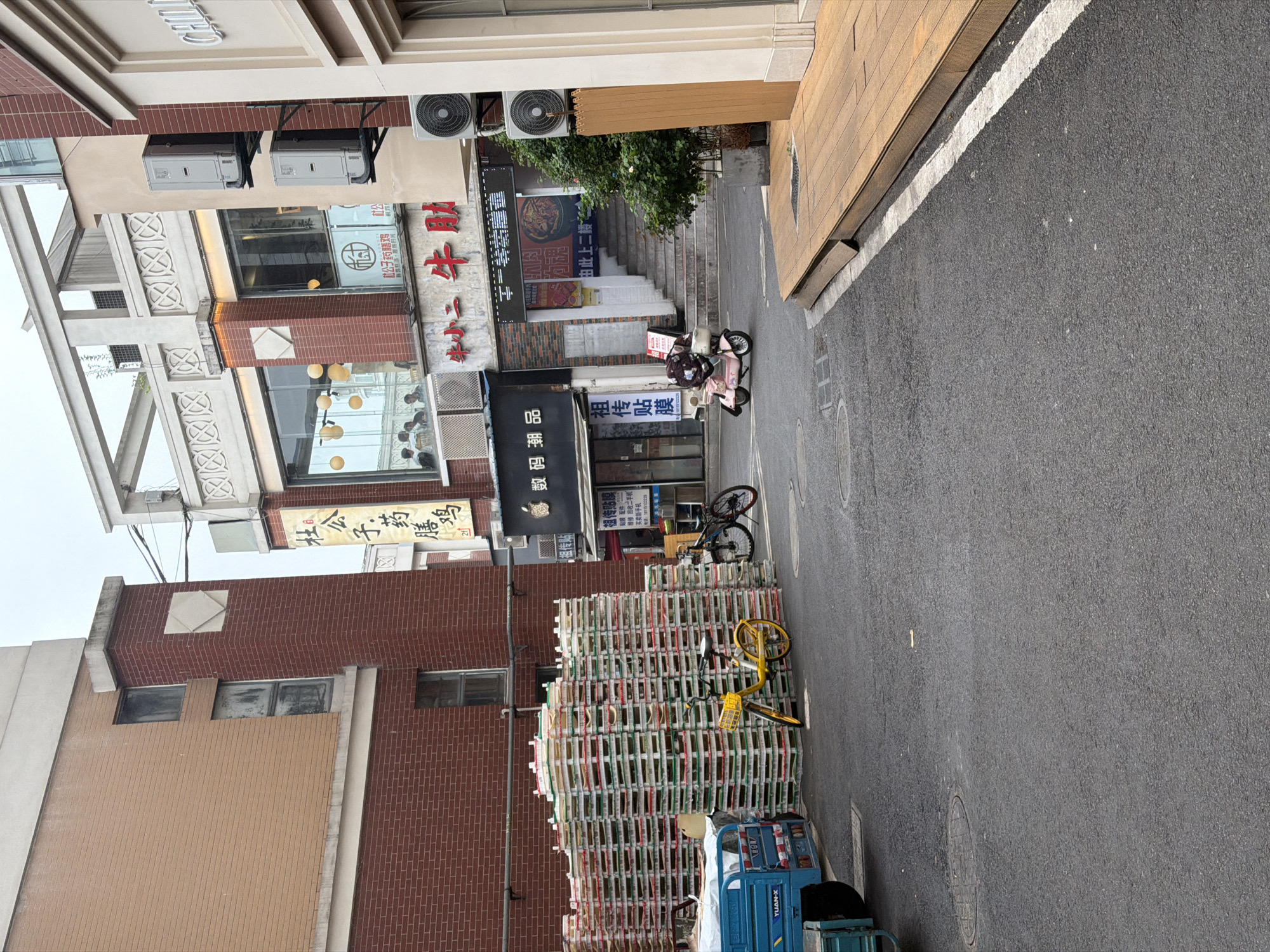 The image captures a street scene in Jiangning District, China. On the left, the facades of buildings feature a mix of brick and stucco, adorned with Chinese signage. A stack of wooden pallets is stacked on the street, next to a three-wheeled blue delivery vehicle. Two bicycles are leaning against the stack of pallets. A person on a pink scooter is positioned in the middle of the street.