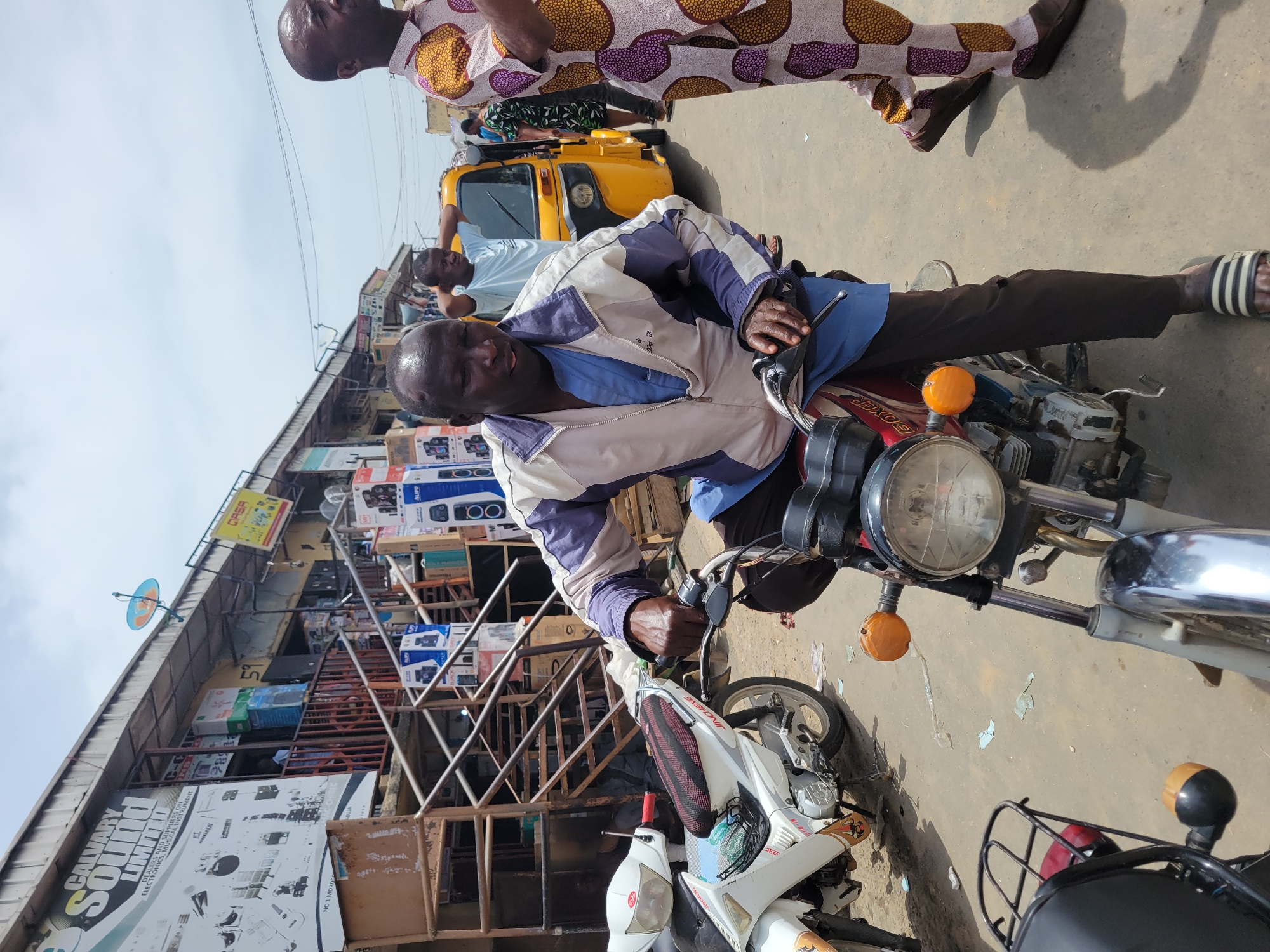 The image depicts a man sitting on a motorcycle in what appears to be a busy street in Suleja, Nigeria. The background features a building with electronics and musical instrument stores like "Calvary Sound Limited." In the foreground, a yellow tuk-tuk and several other motorcycles are visible. A man in a patterned outfit stands nearby. The overall scene suggests a bustling commercial area.